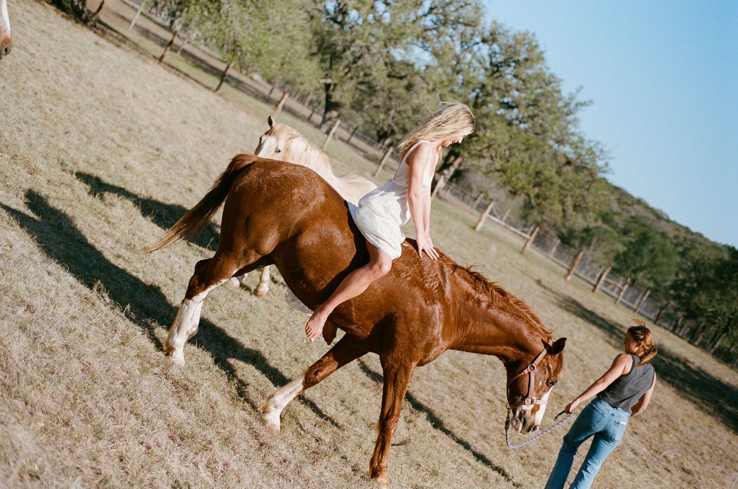 Rider on a bucking chestnut horse in an open field with a handler nearby.