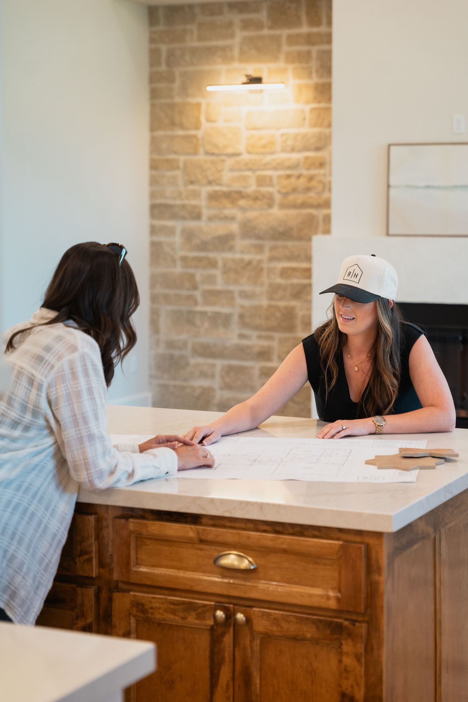 Two women reviewing blueprints on a marble kitchen island in a stone-accented home.