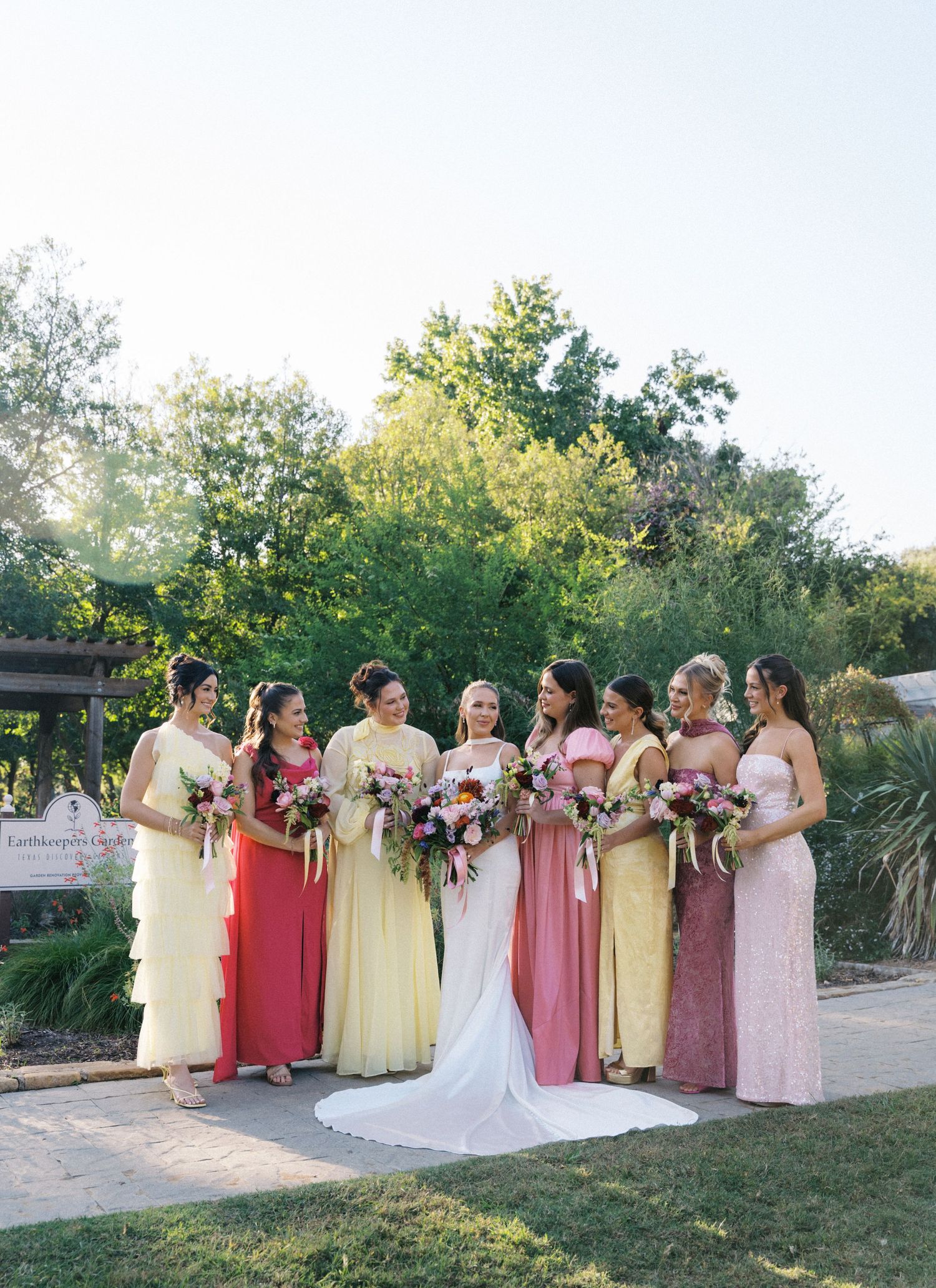 Bride in white gown poses with seven bridesmaids in mismatched yellow, coral, and pink gowns holding bouquets.