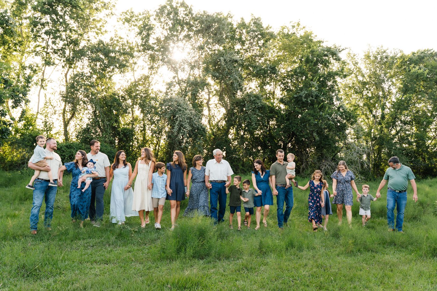Large extended family walking together in a lush green field surrounded by trees during golden hour outdoor photo session.