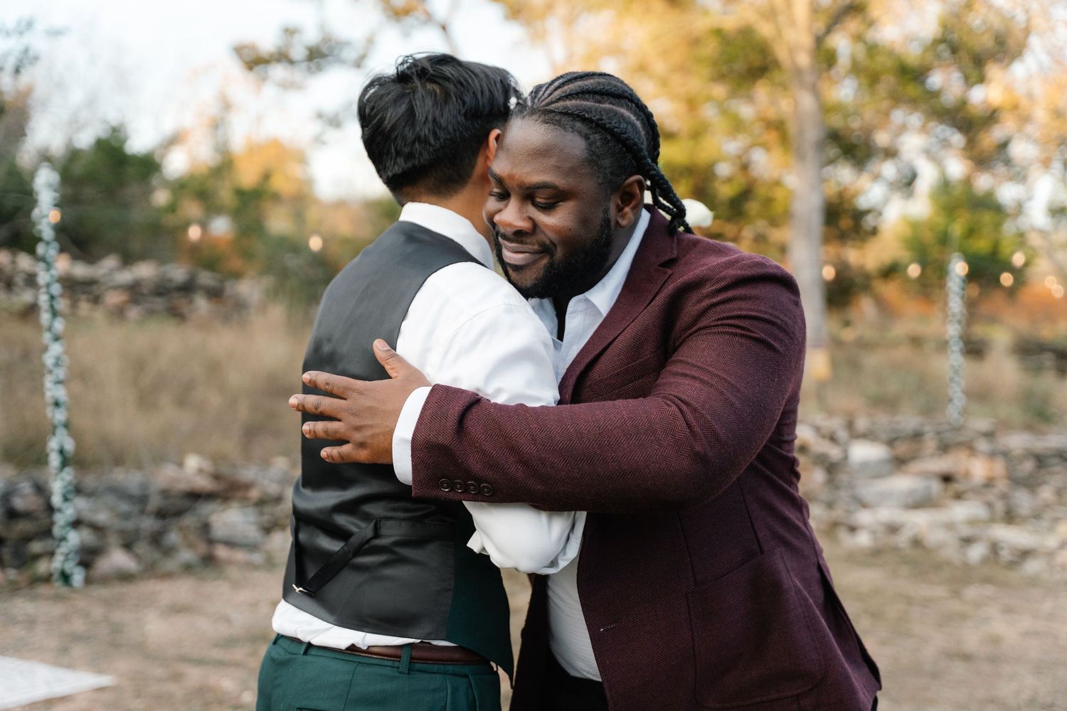 Two men sharing a warm hug in formal attire at outdoor wedding ceremony.