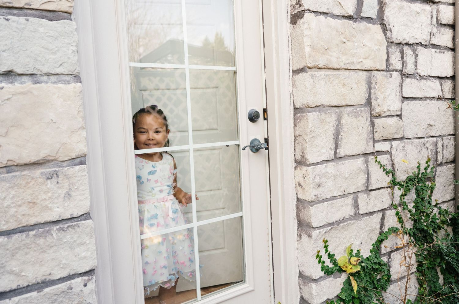 Adorable toddler in floral dress peeking through glass door at wedding venue.