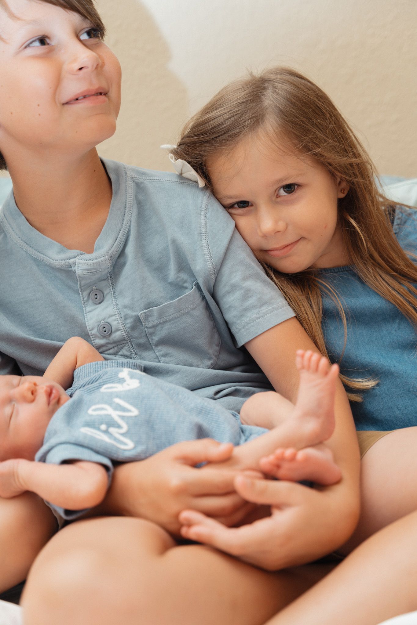 Two older siblings in blue outfits tenderly hold their newborn baby brother.