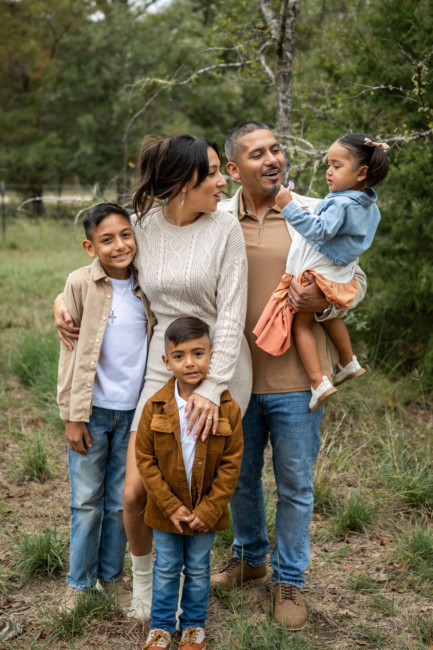 A happy Hispanic family of five poses together outdoors in coordinating fall outfits among trees and green grass.