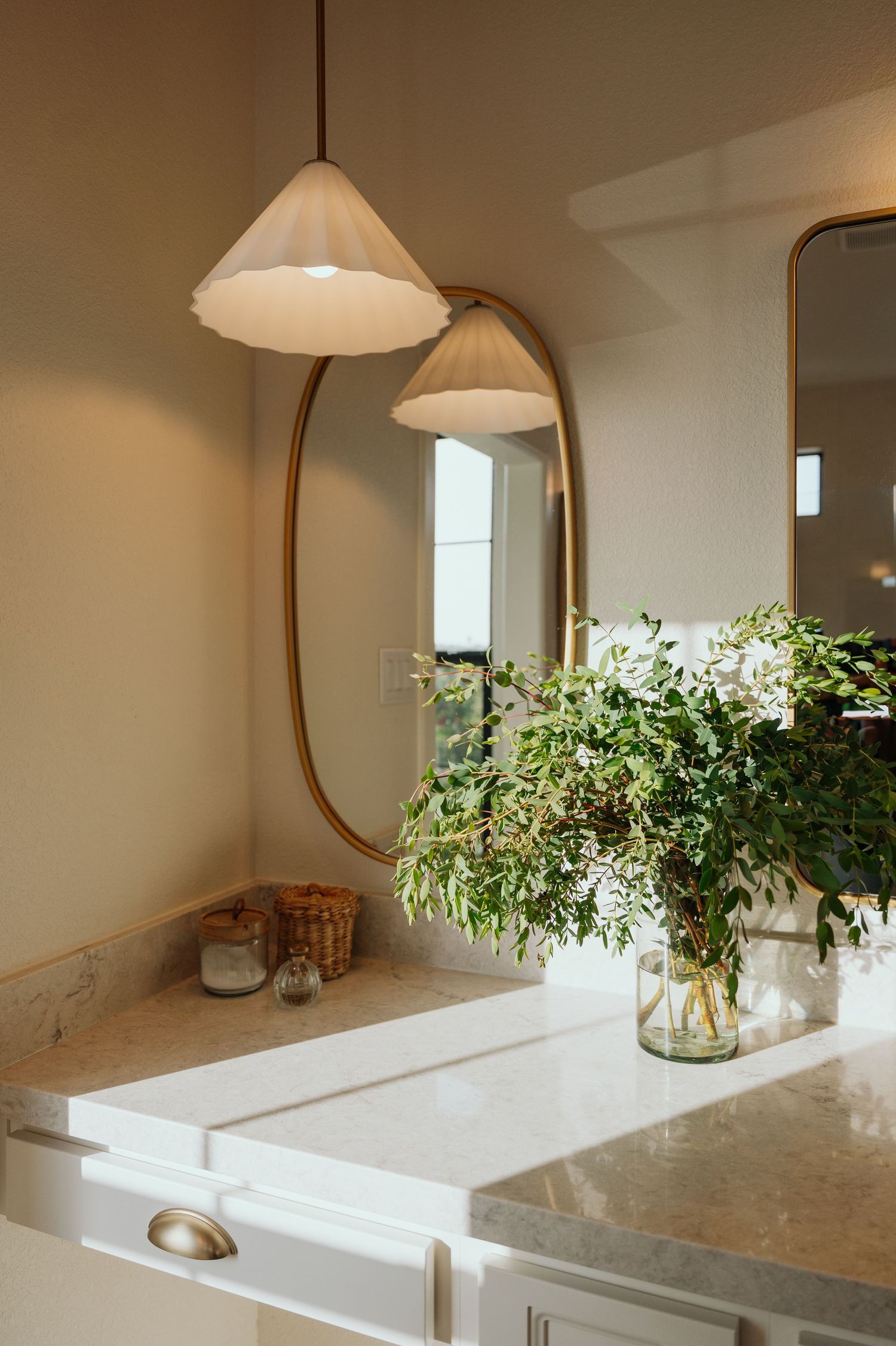 Elegant bathroom vanity with oval gold mirror, pendant light, marble countertop, and fresh greenery arrangement.
