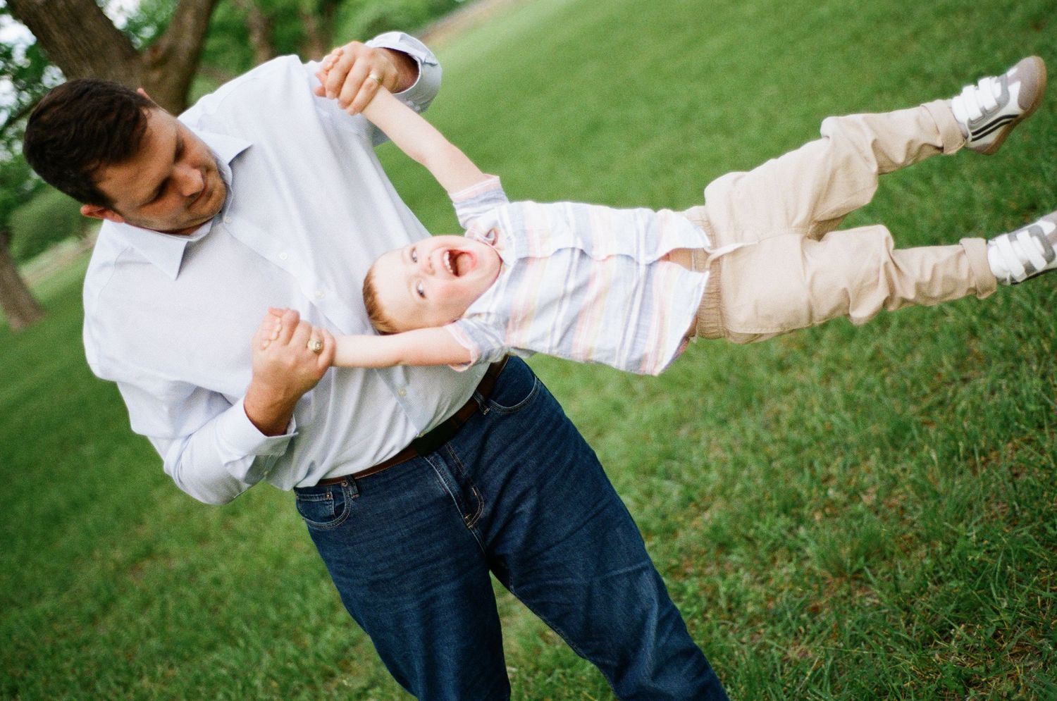 A father playfully swings his laughing young son in a grassy outdoor setting.