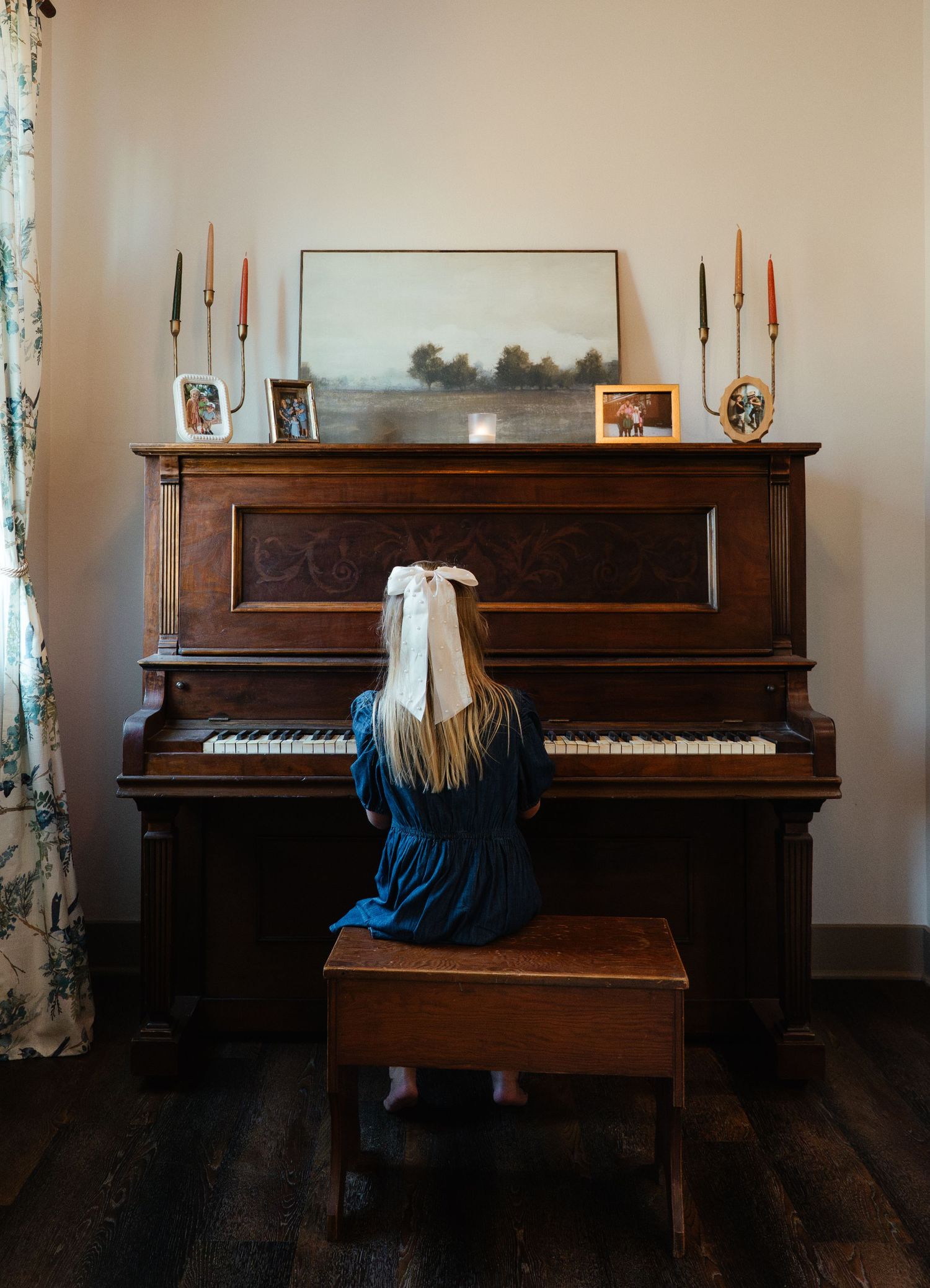 A young girl in a blue dress plays an antique upright piano indoors.