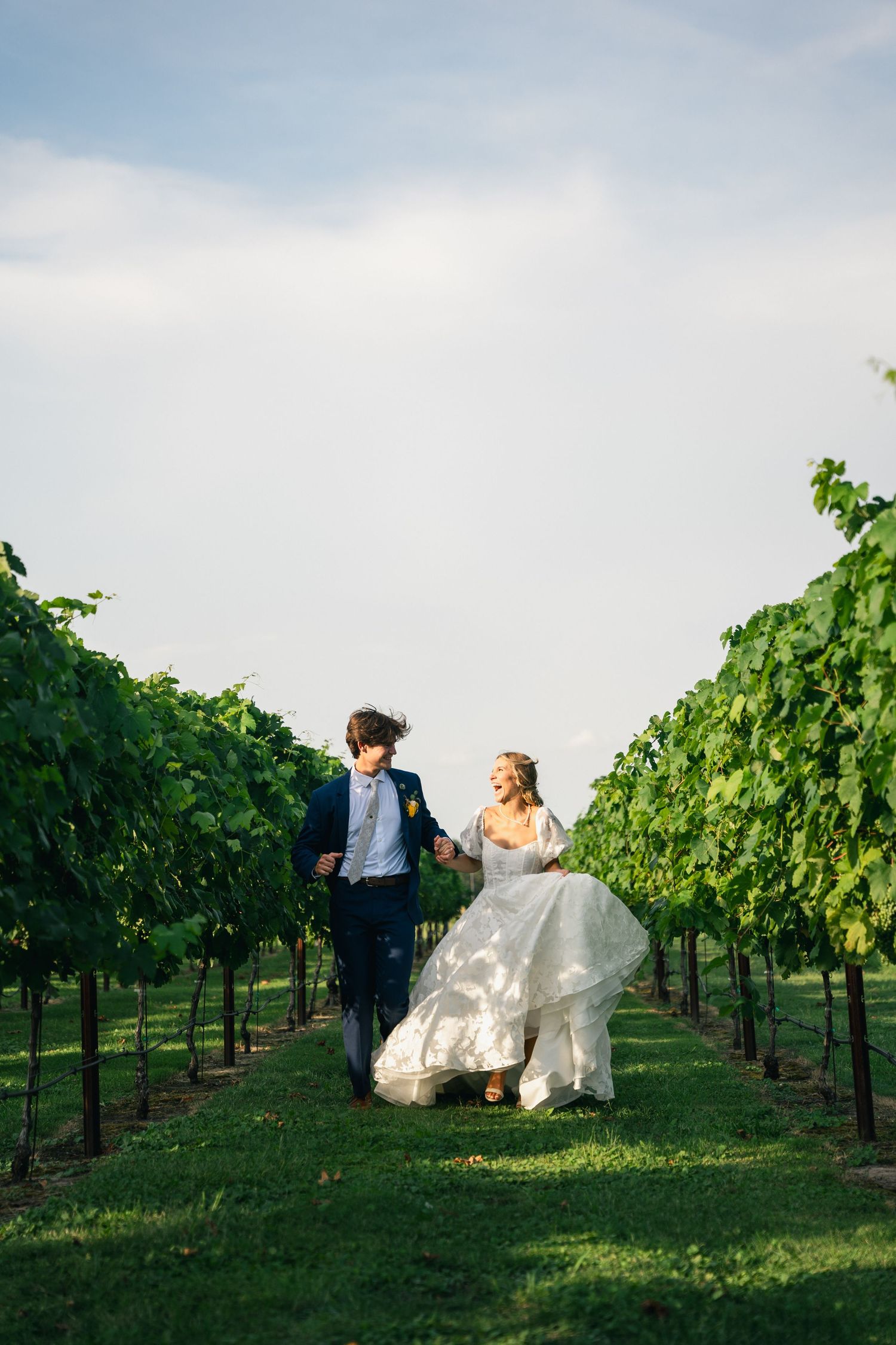 Bride and groom holding hands running through lush green vineyard rows on their wedding day.