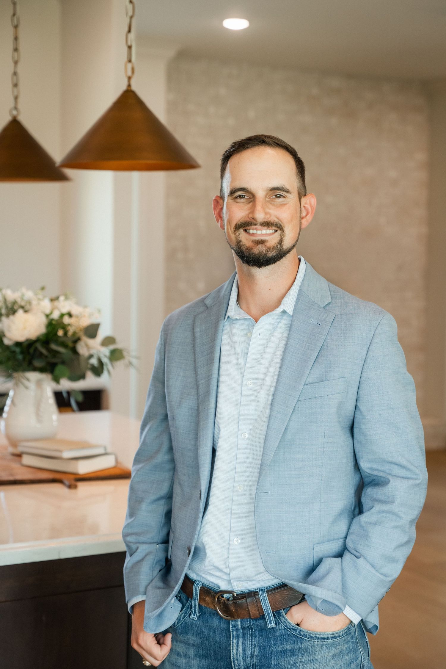 Smiling man in light blue blazer and jeans standing in modern home interior with pendant lights.