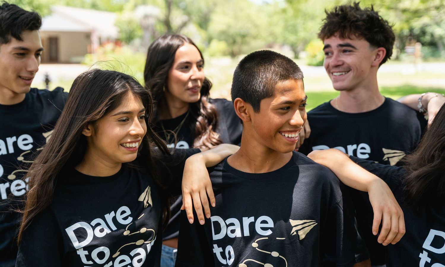 Diverse teens in matching Dare to Dream black t-shirts smiling and embracing outdoors.