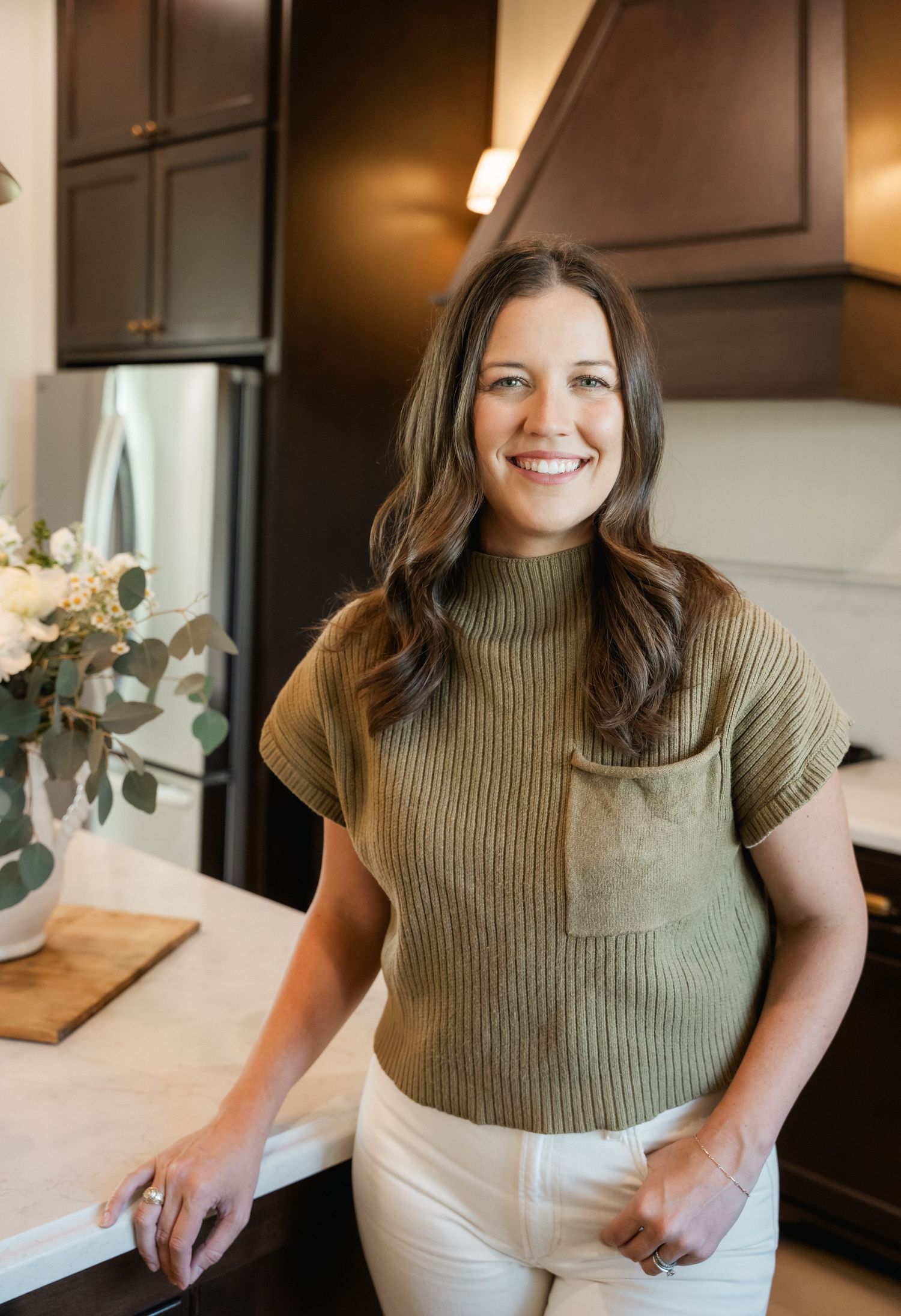 Smiling brunette woman in green ribbed top leaning on white kitchen counter.