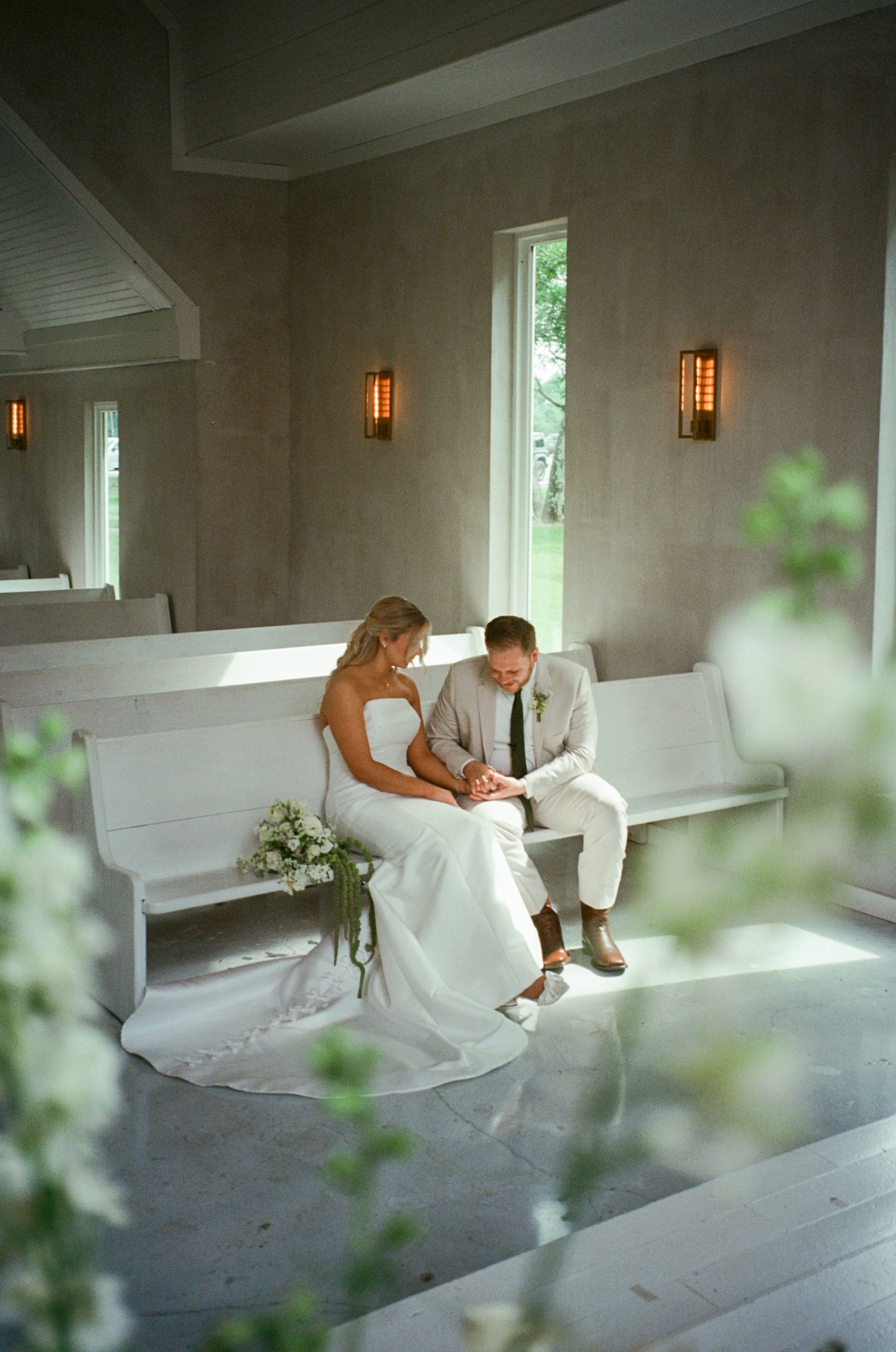Bride and groom sit together on a white chapel pew, sharing a quiet moment in a softly lit wedding venue.