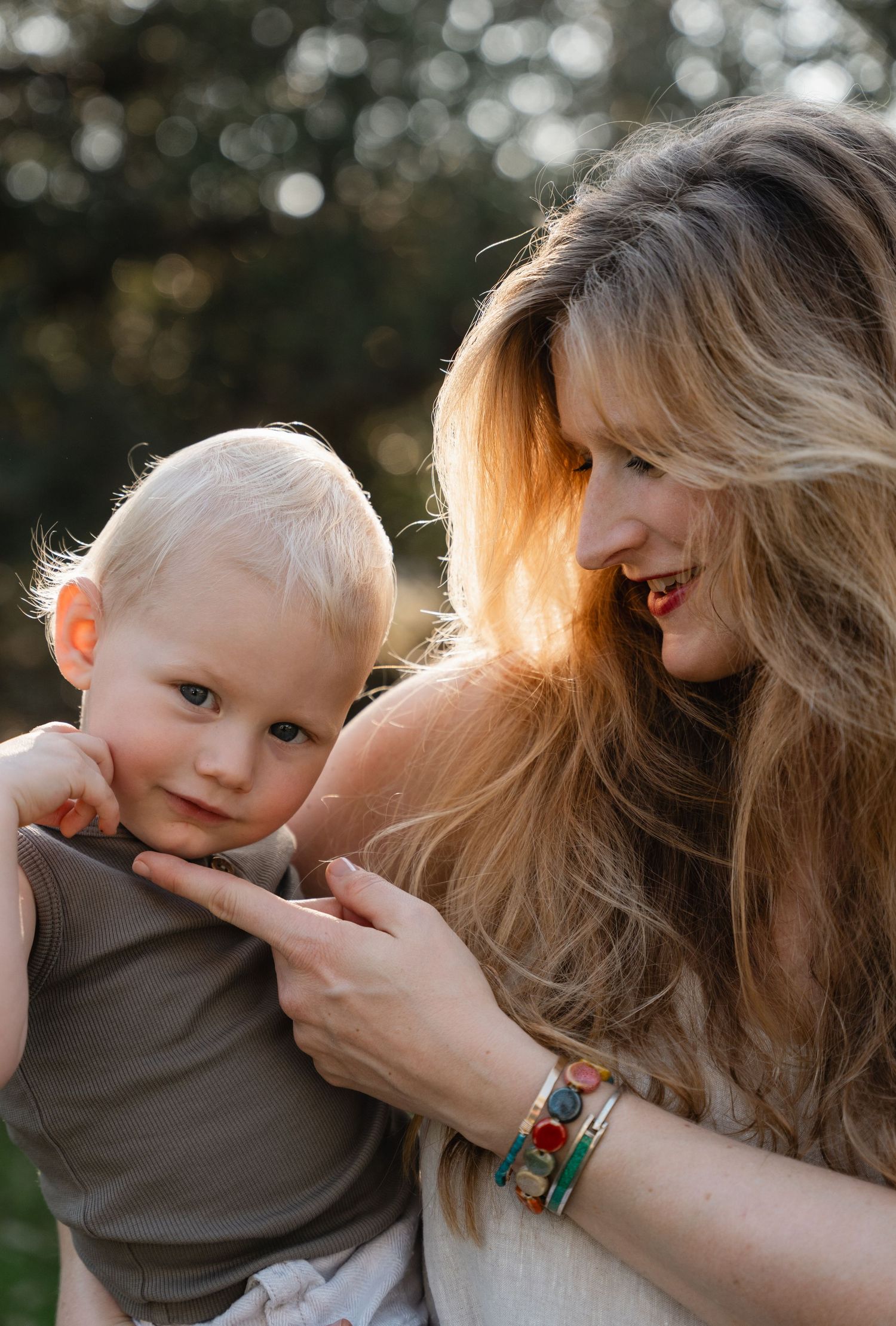 Loving mother with long hair smiling at her blonde baby boy outdoors in golden light.