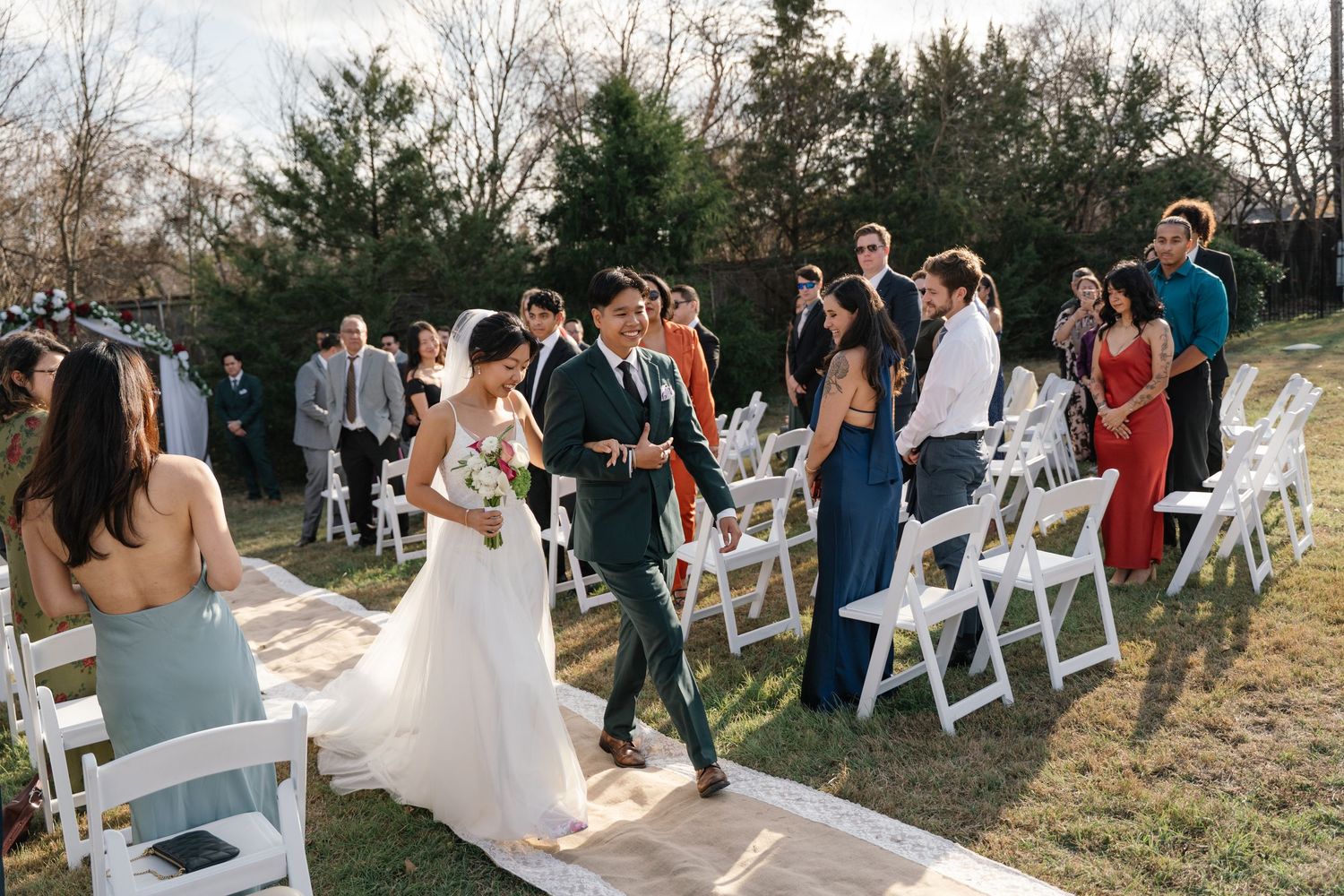Newlyweds walk back down aisle together as guests celebrate at outdoor backyard wedding ceremony.