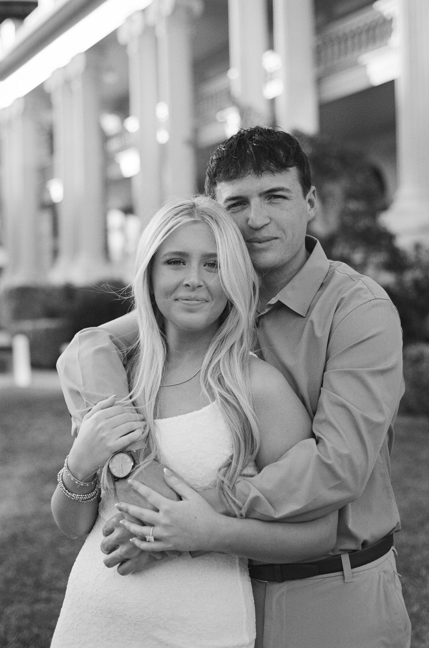 Black and white photo of a smiling couple embracing outdoors, woman wearing white dress and ring, man in button-up shirt.