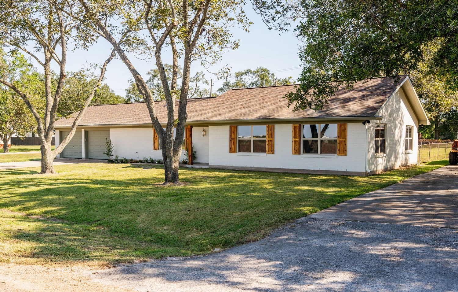 White ranch-style home with brown shutters, large trees, and a well-maintained green lawn and gravel driveway.