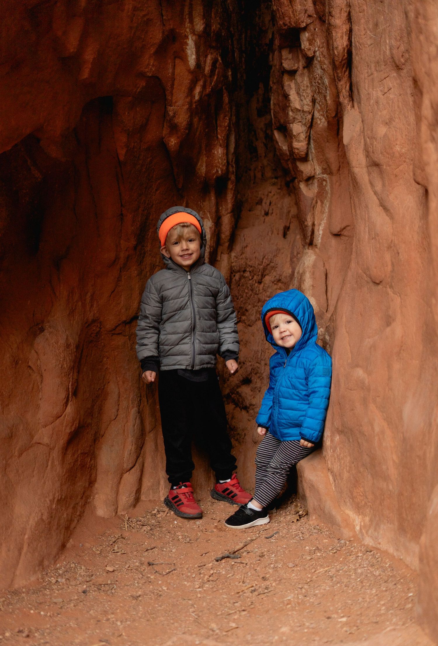 Two children in colorful winter jackets explore a red rock canyon slot cave during an outdoor hiking adventure.