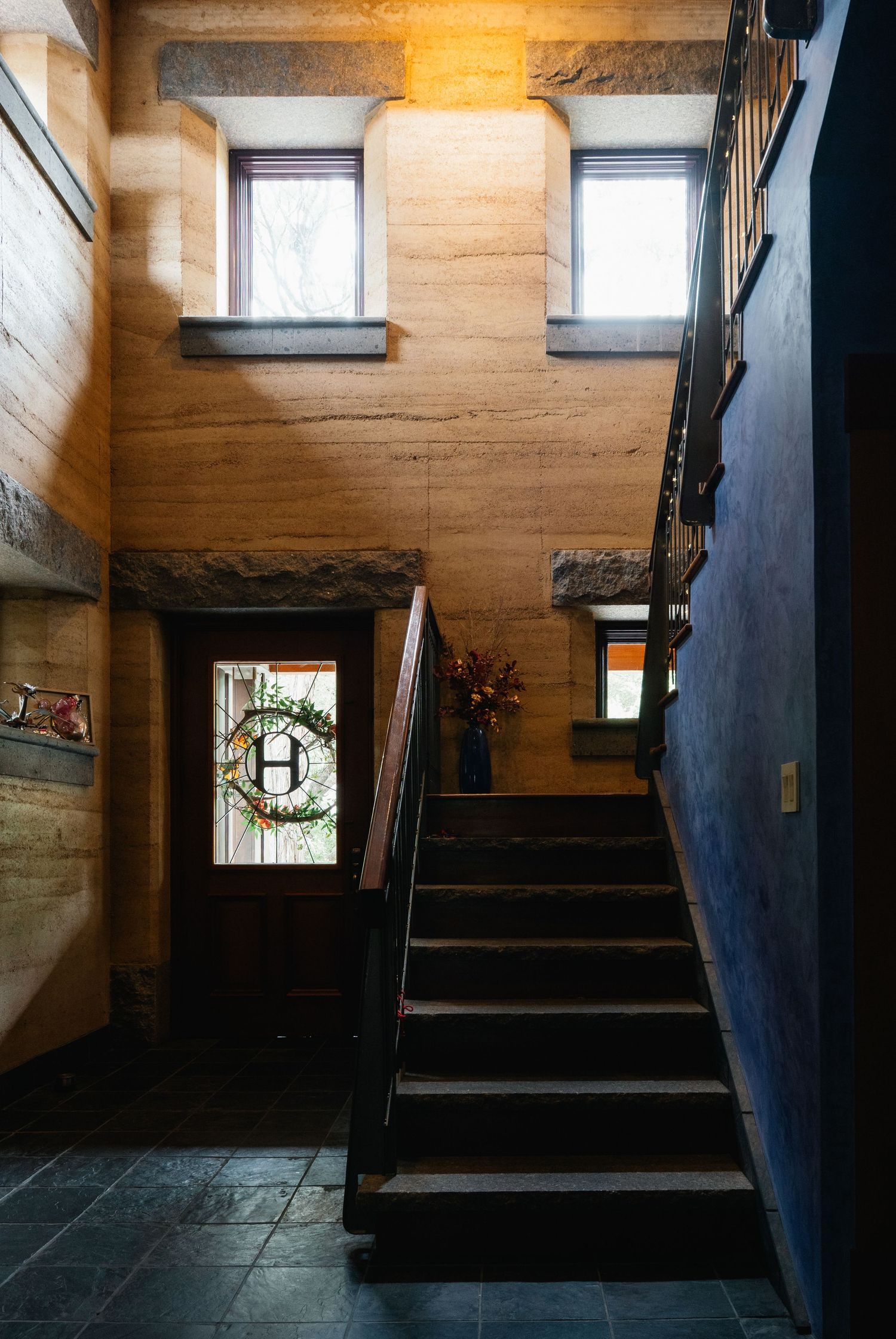 Rustic stone entryway with dark wood staircase, wreath-decorated front door, and high windows in warm lighting.
