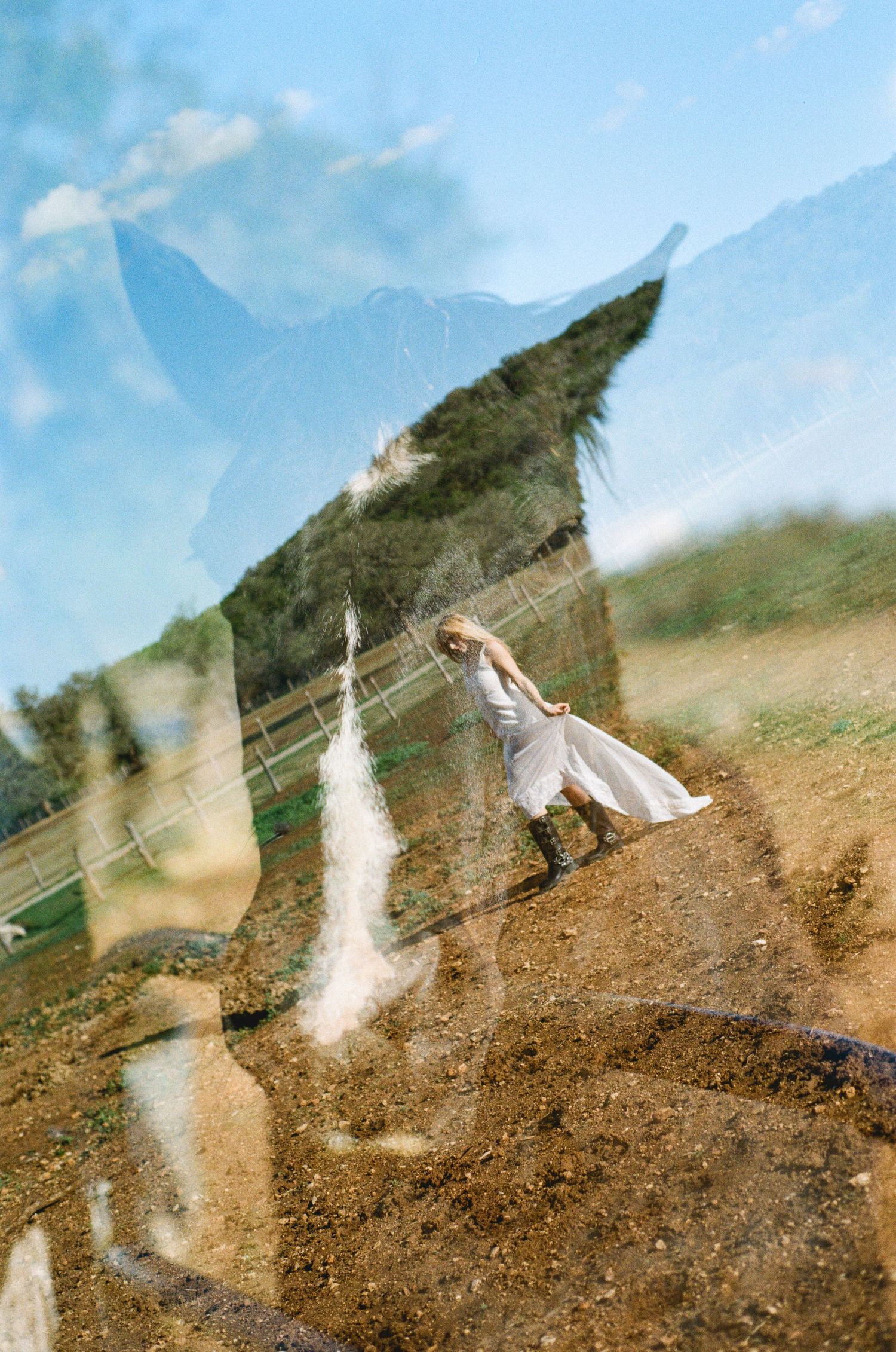 Double exposure photo of a woman in white dress on a farm with mountain landscape.