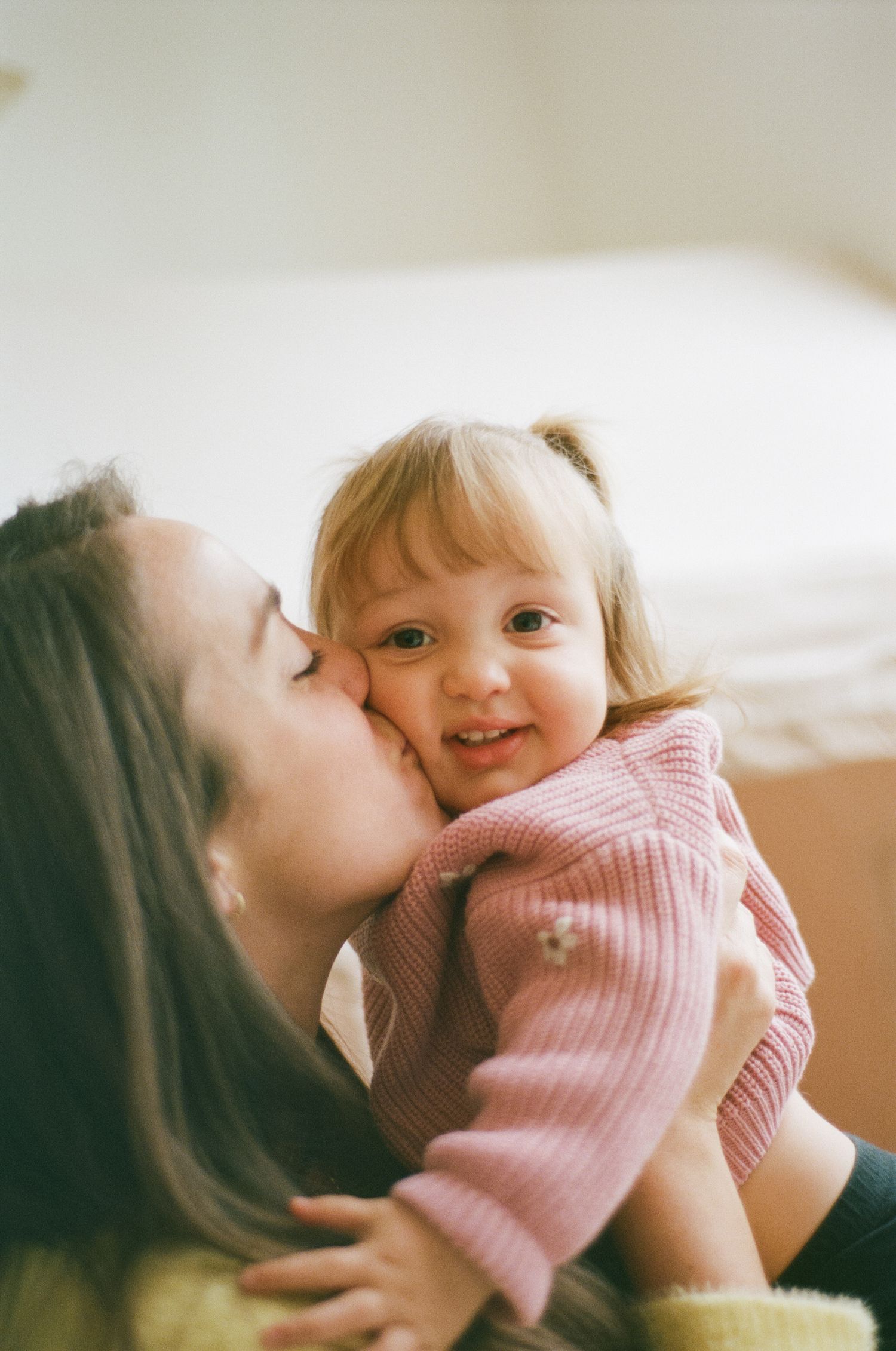 A mother tenderly kisses her smiling toddler girl in a pink sweater on the cheek.