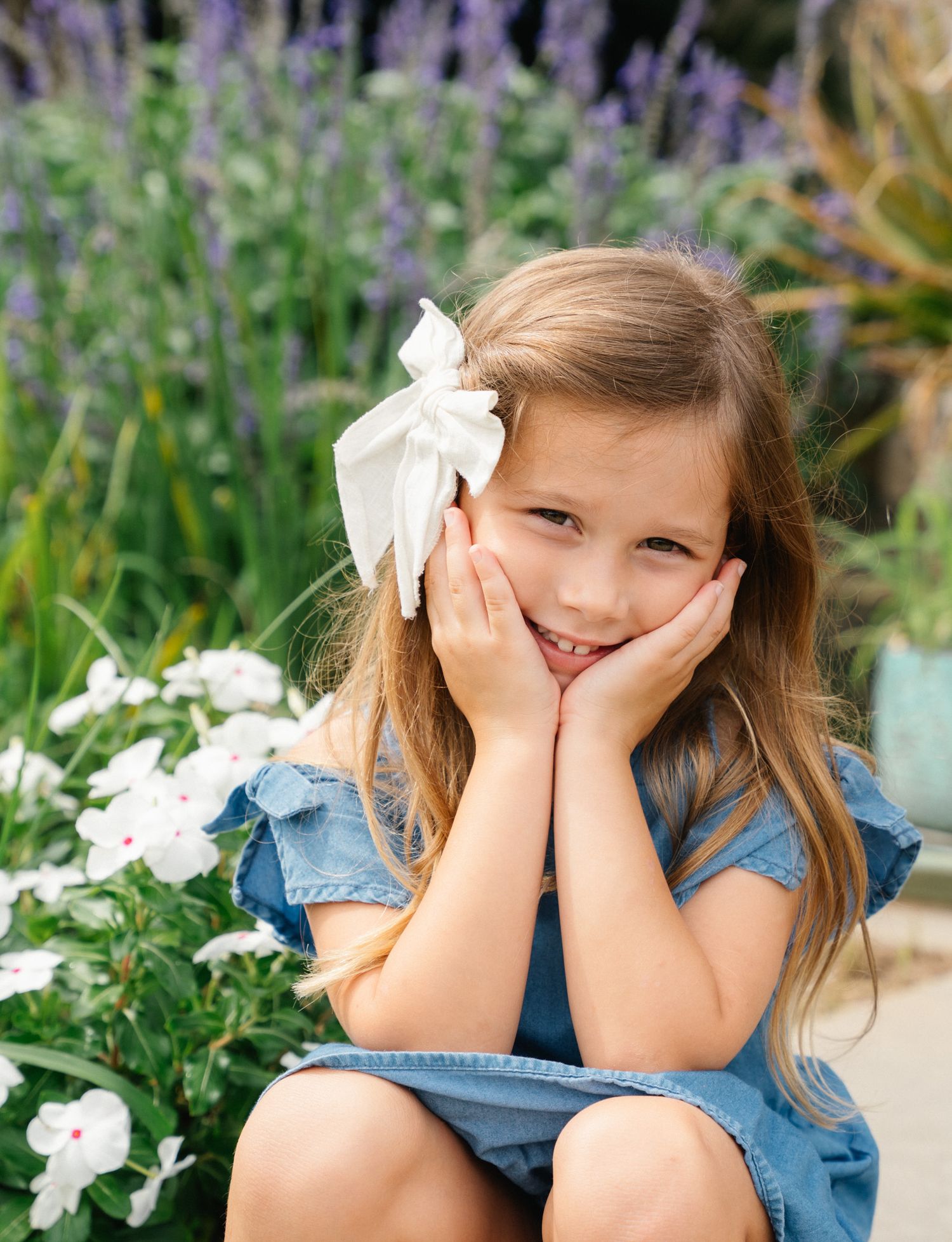 A cute young girl in a denim dress with a white bow rests her cheeks in her hands among purple garden flowers.