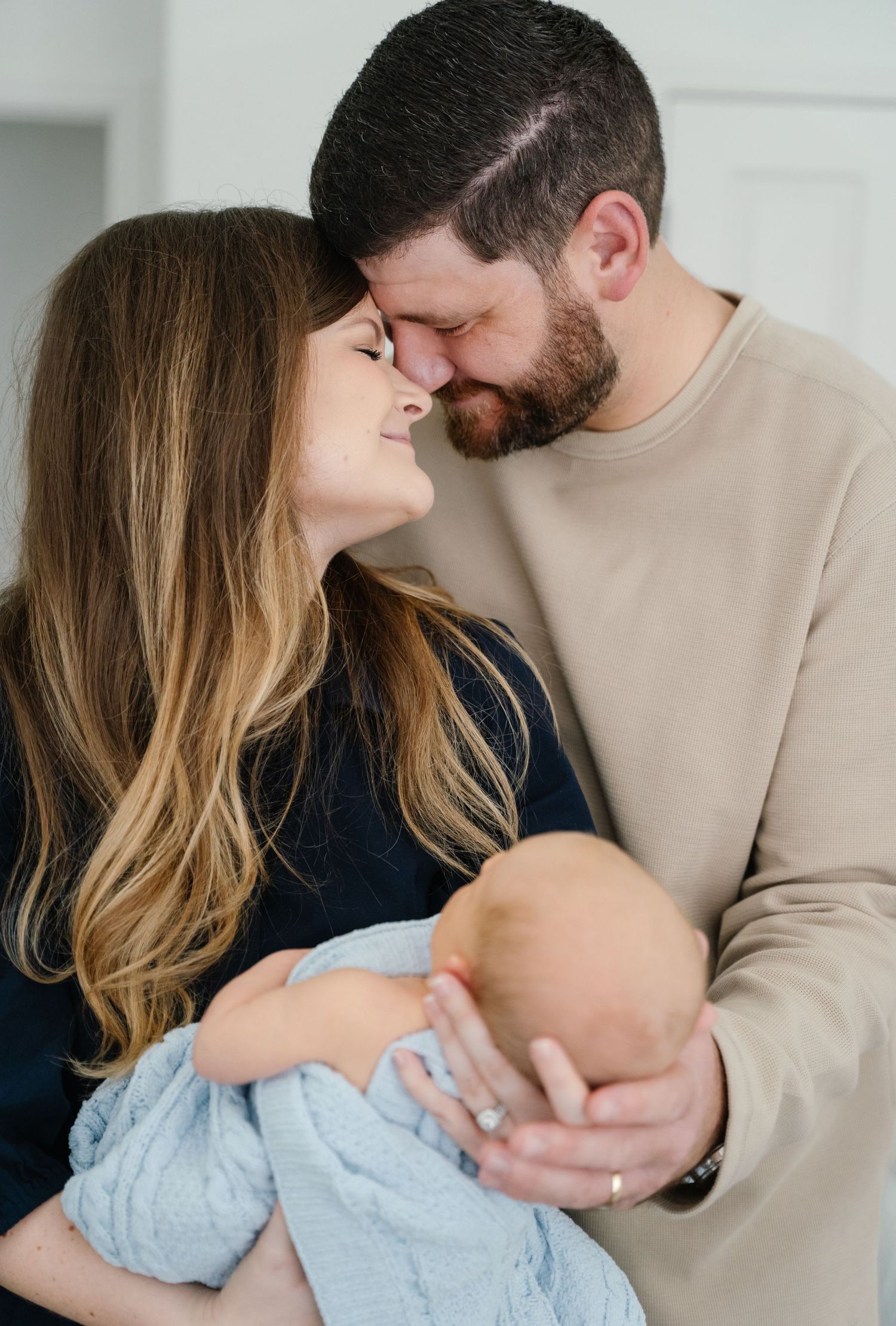 New parents touching foreheads tenderly while holding newborn baby during intimate home session.