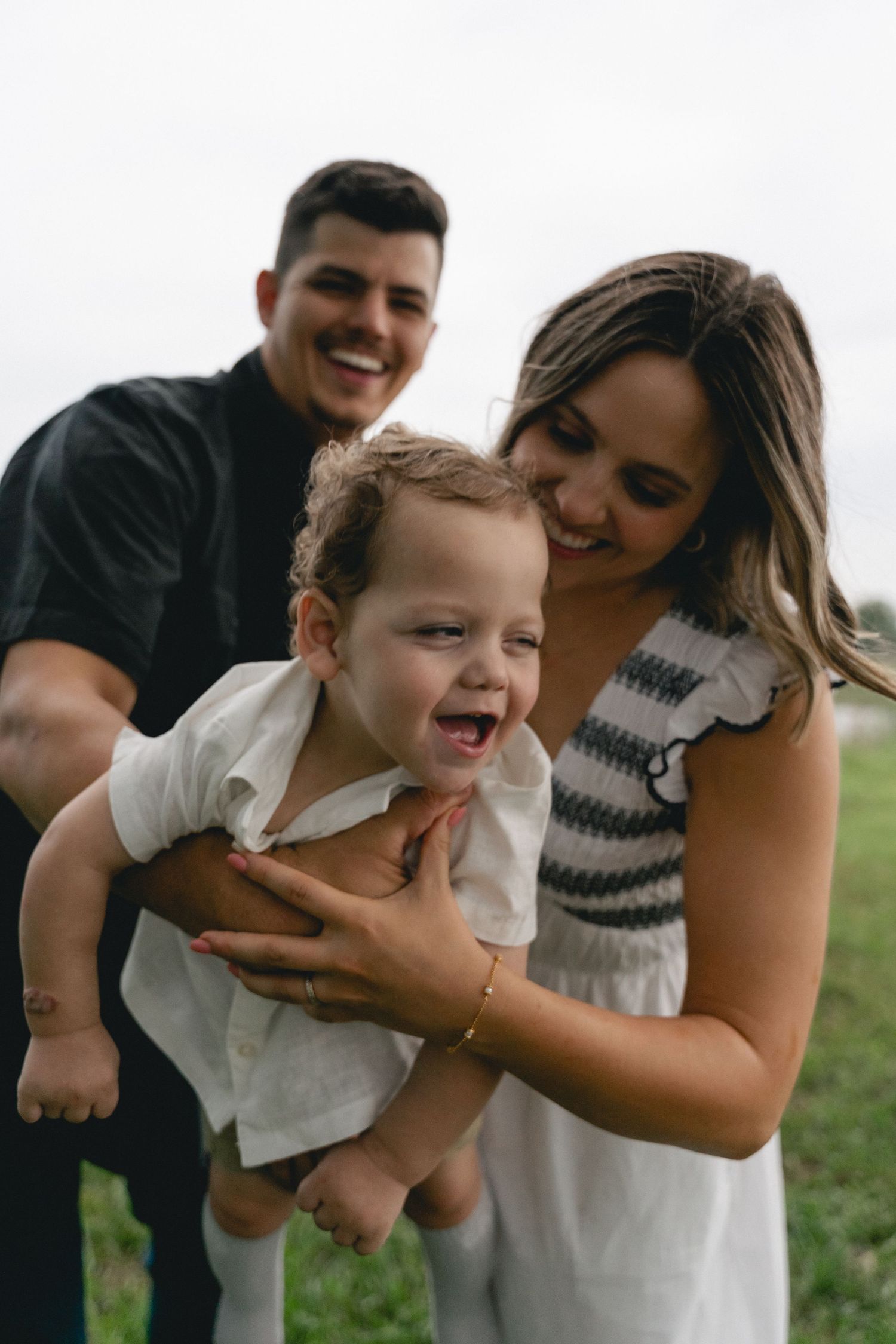 Happy young family laughing outdoors in a green field, parents holding their giggling baby between them.