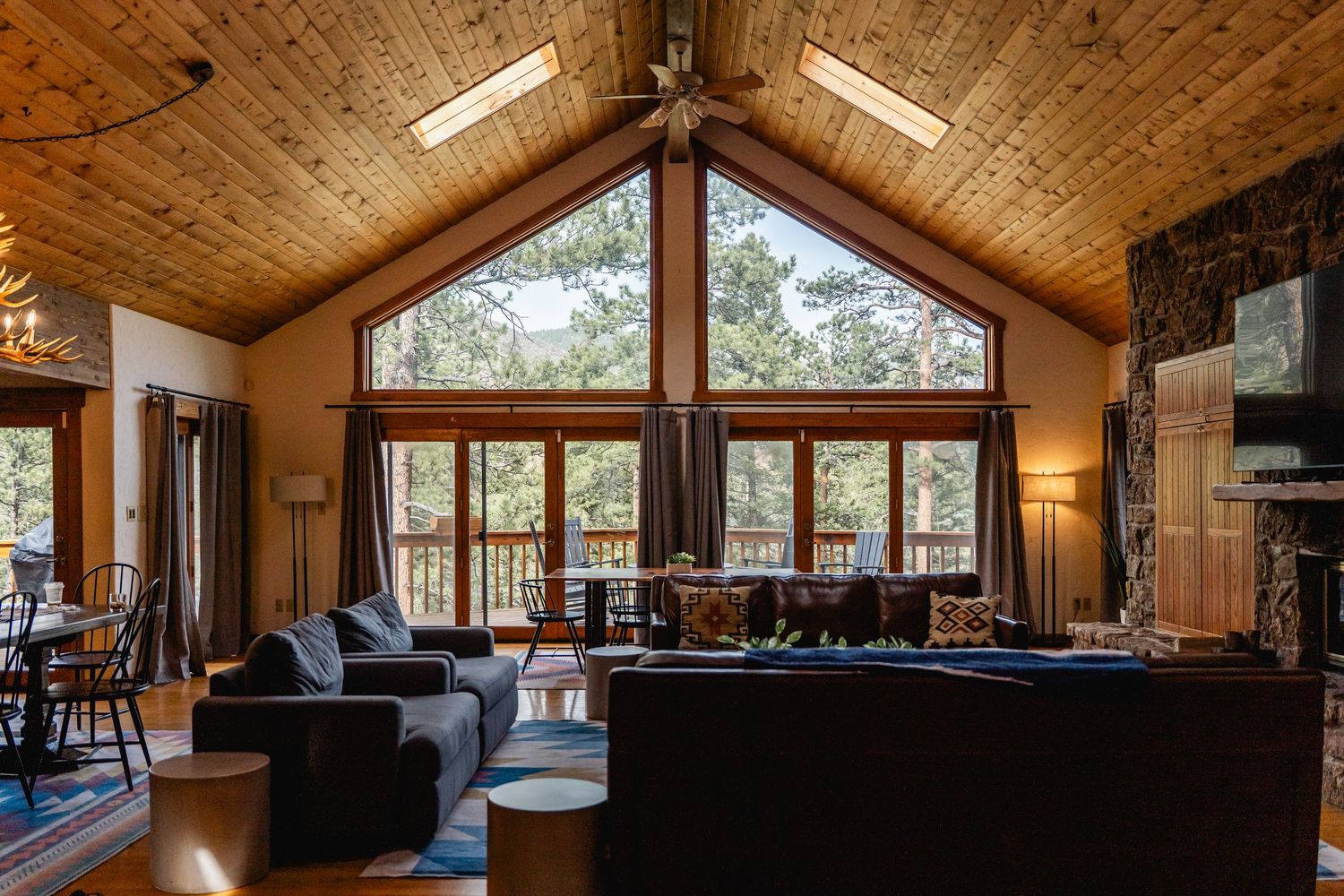 Spacious log cabin living room with vaulted wood ceiling, large windows overlooking forest, and stone fireplace.