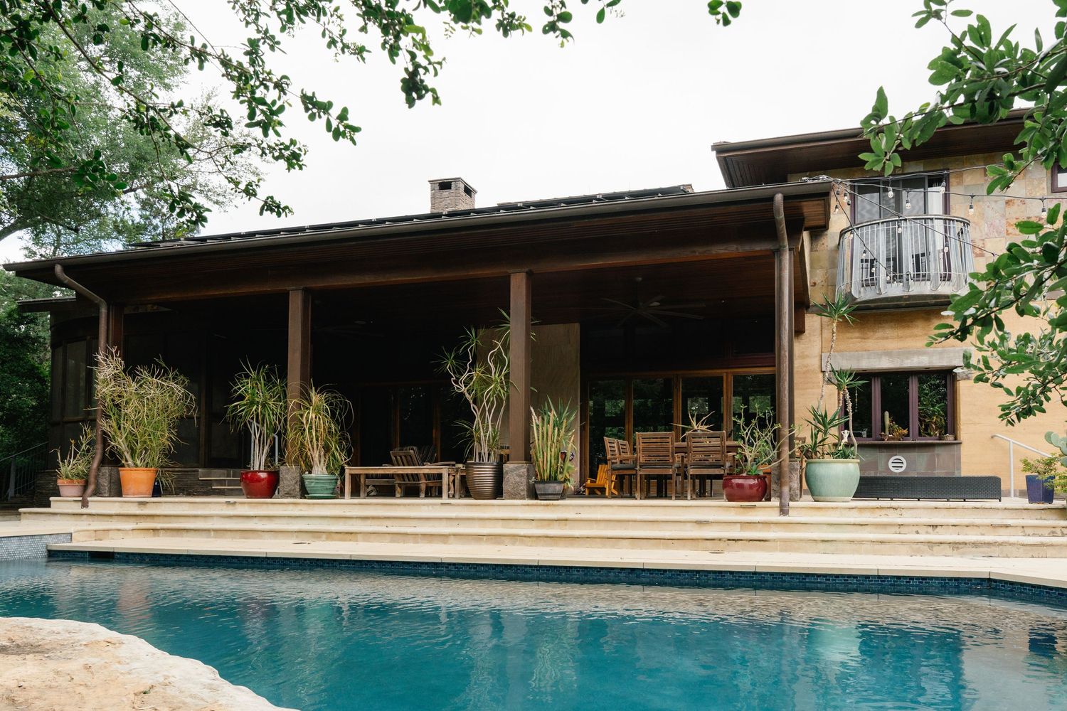 Luxury home exterior with covered patio, potted plants, outdoor dining, and sparkling swimming pool in foreground.