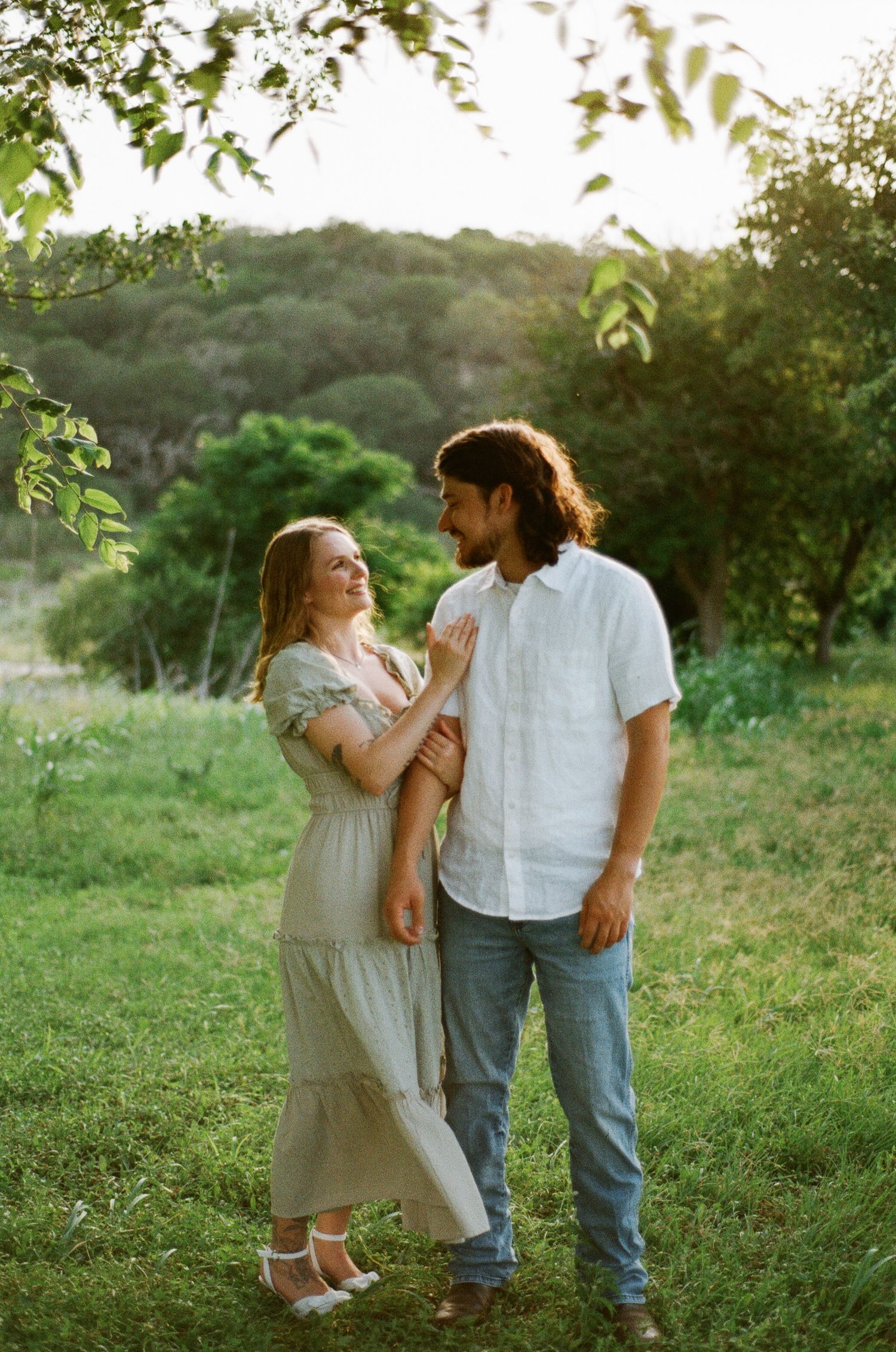 Couple standing in a sunlit green meadow, woman in flowy dress smiling at bearded man in white shirt and jeans.