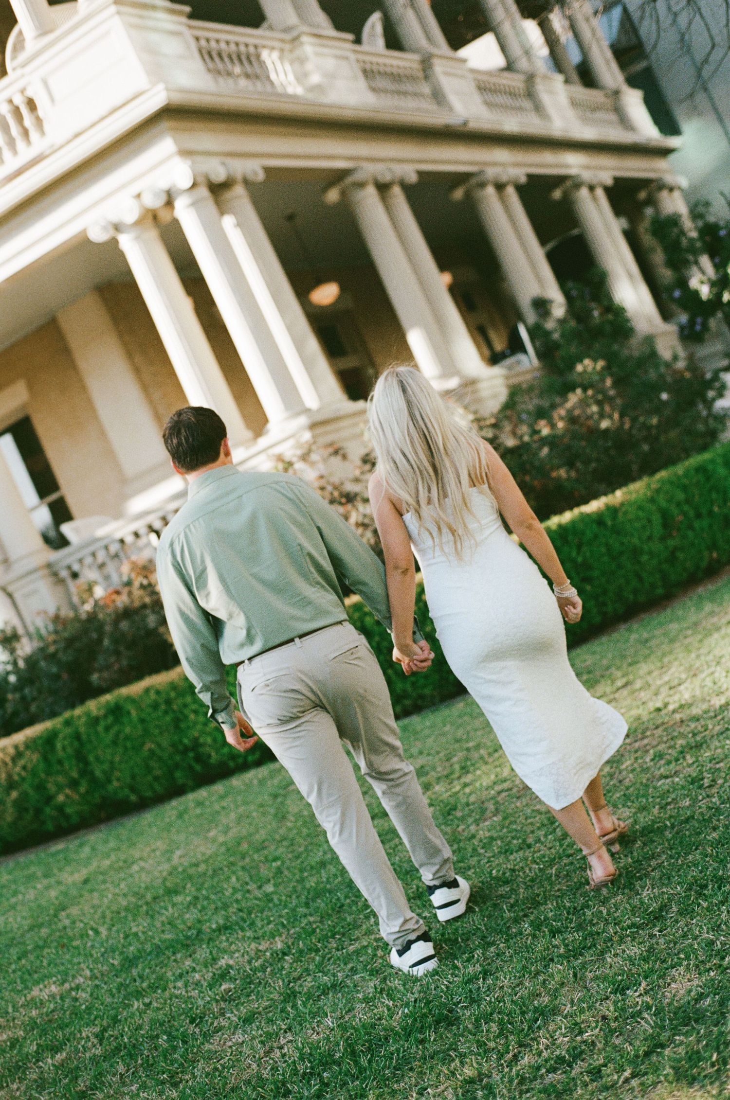 Couple holding hands walking toward a grand white mansion with tall columns on a lush green lawn.