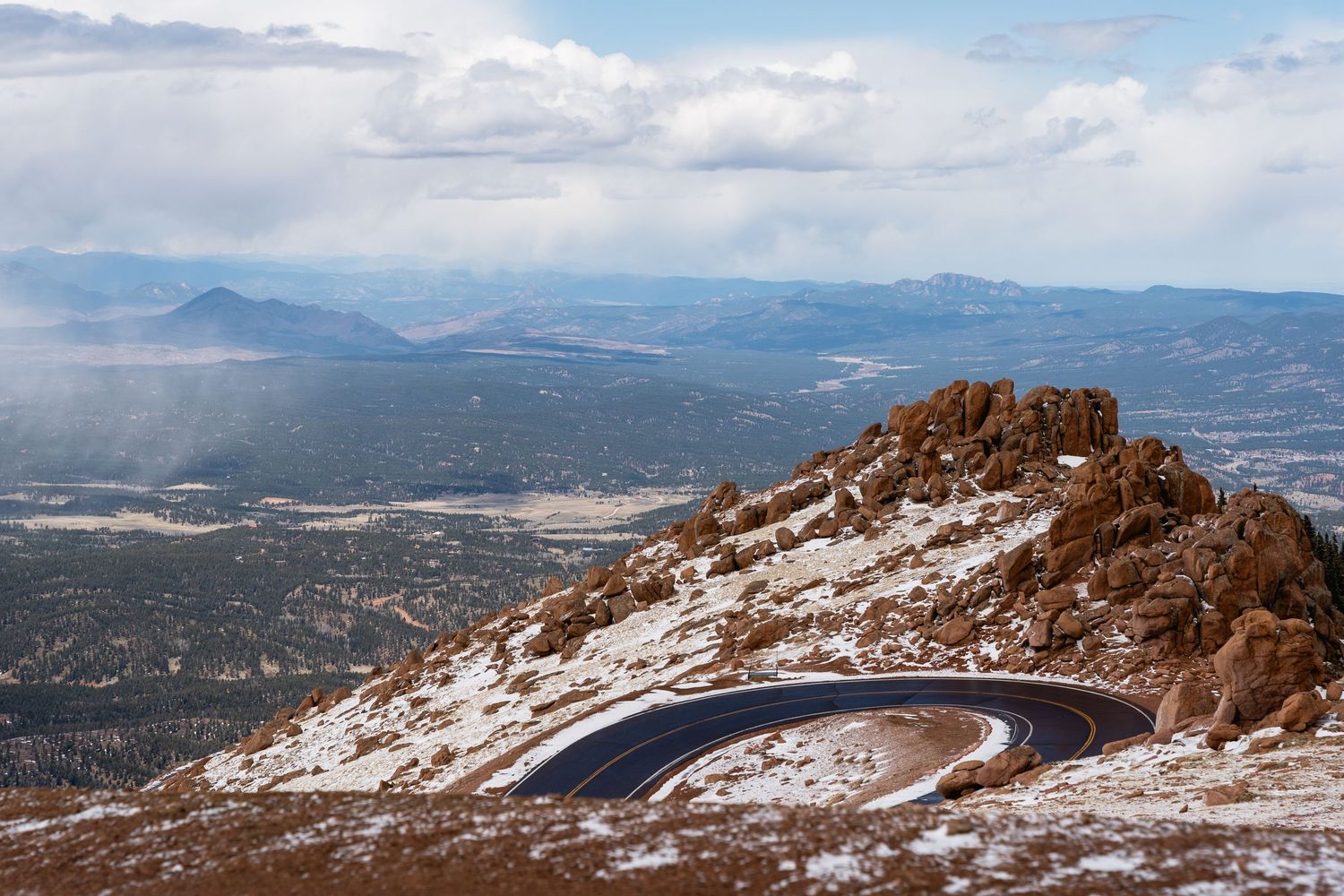 Aerial view of winding Pikes Peak Highway switchback road with snow-dusted Rocky Mountain landscape below.