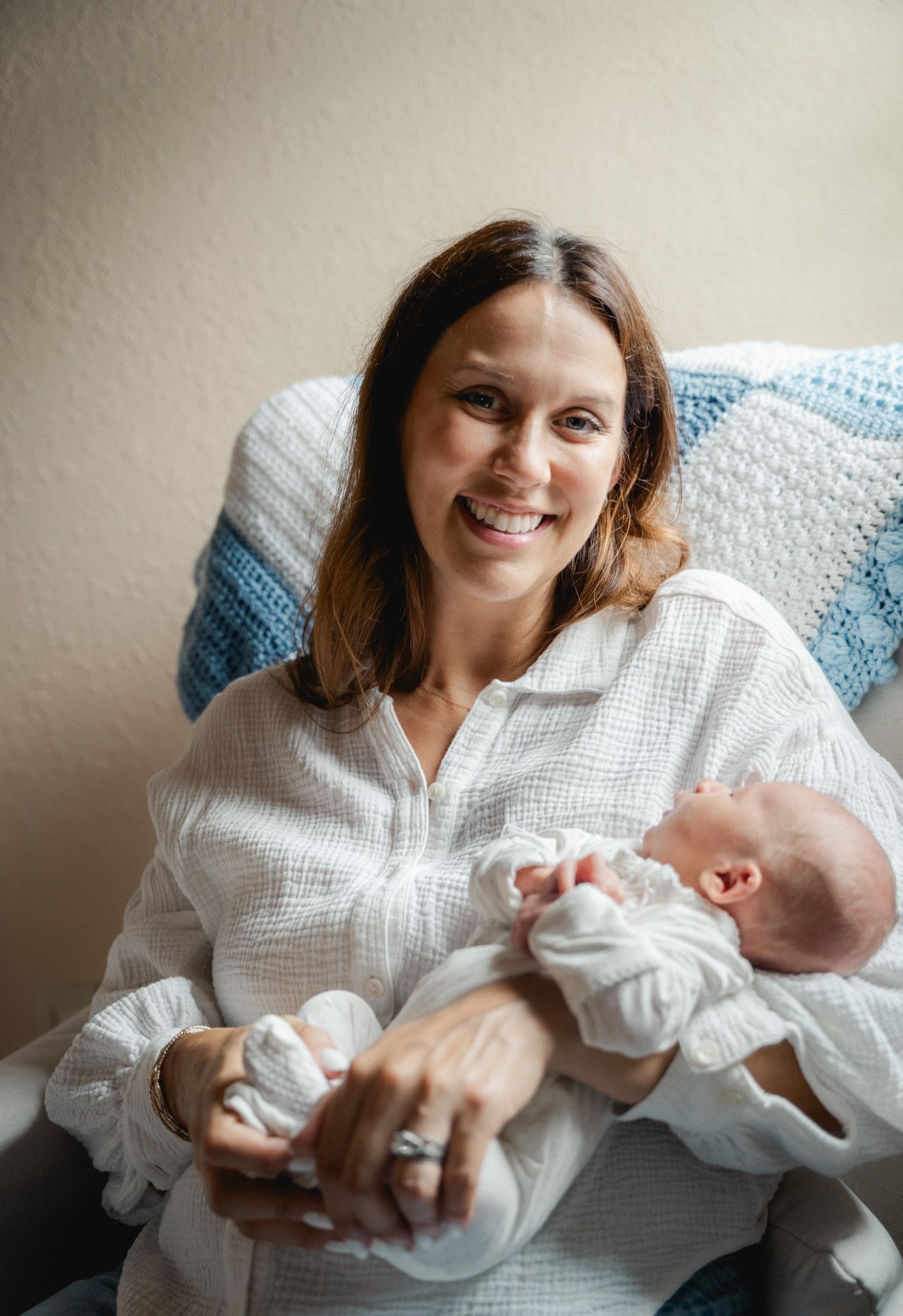 Smiling brunette mother in white shirt holding newborn baby in nursery chair.