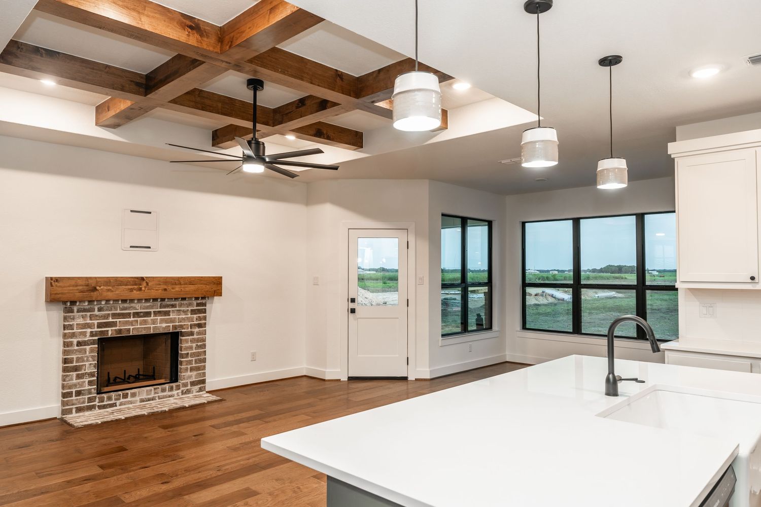 Open living area with coffered ceiling, brick fireplace, kitchen island, and hardwood floors.