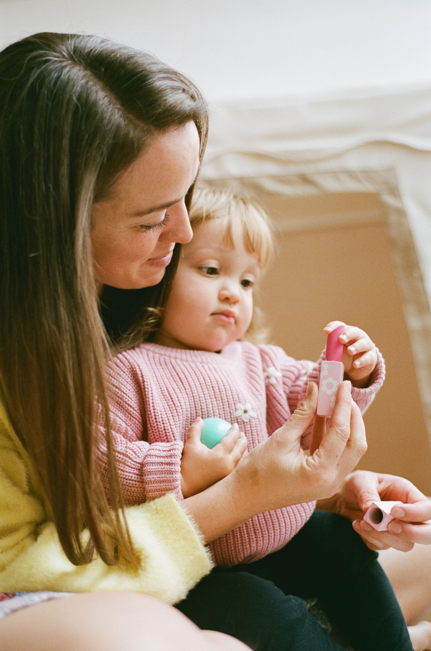 A mother sits with her toddler on her lap, showing her a pink toy in soft natural light.