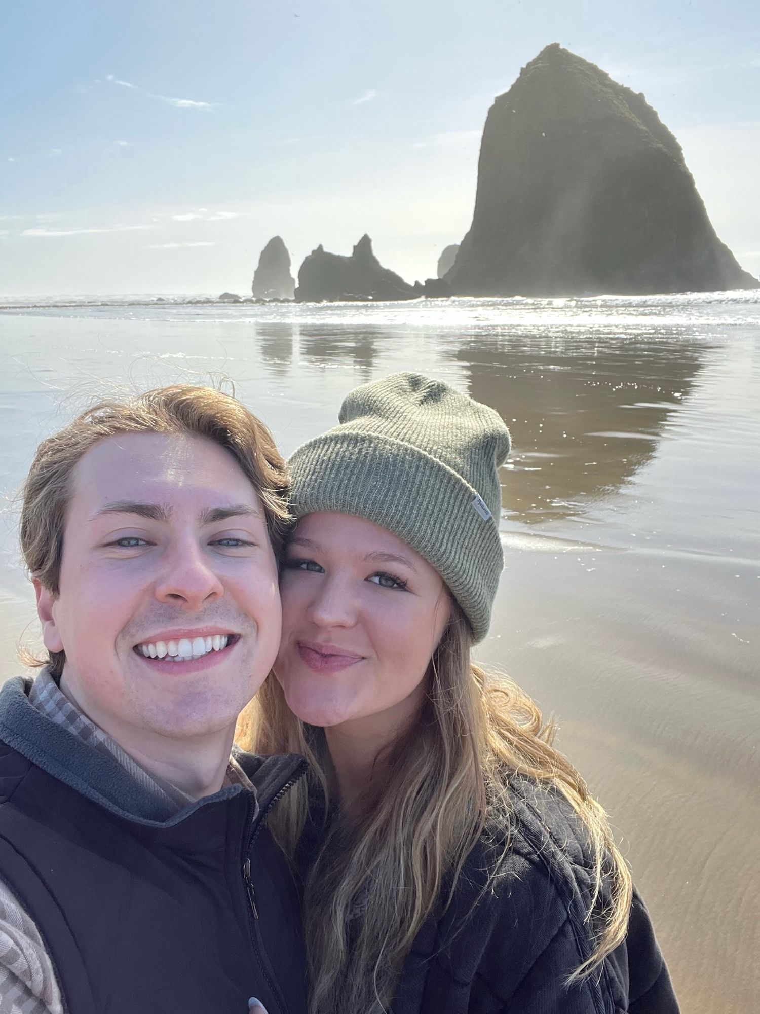 Smiling couple taking a selfie at Cannon Beach with iconic Haystack Rock behind them.