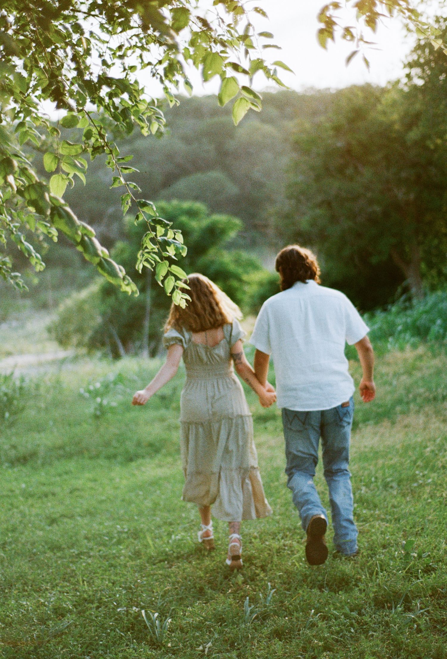 A couple holds hands while walking through a lush green meadow surrounded by trees in warm golden light.