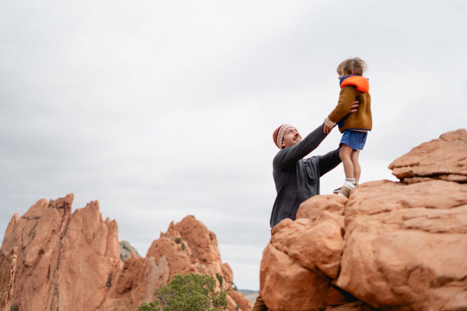 An adult lifts a young child onto a red rock formation at Garden of the Gods under an overcast sky.