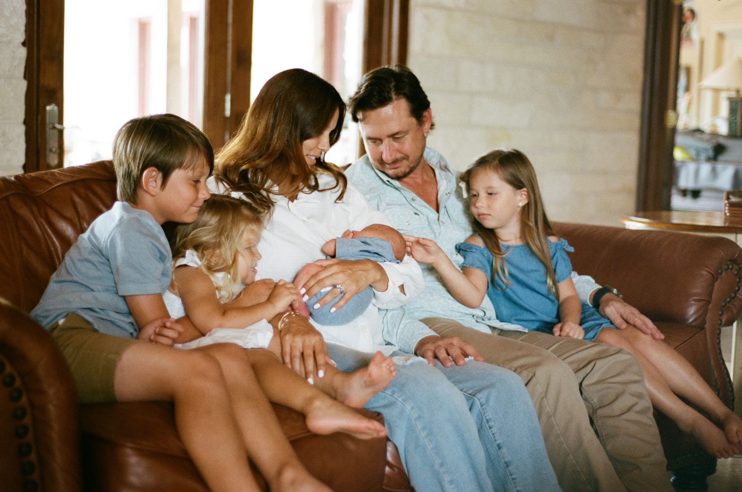 Family of five with newborn sitting together on brown leather couch.