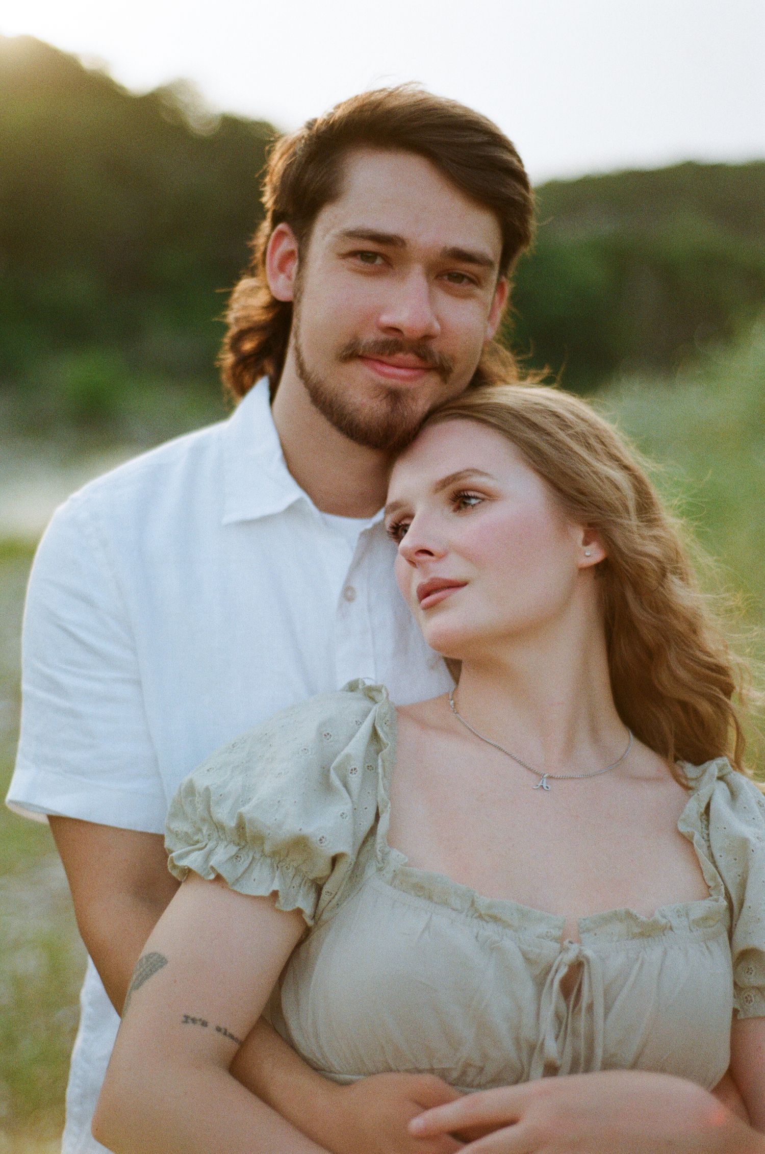 Couple embracing at golden hour, man in white shirt holds woman in sage ruffle dress outdoors.