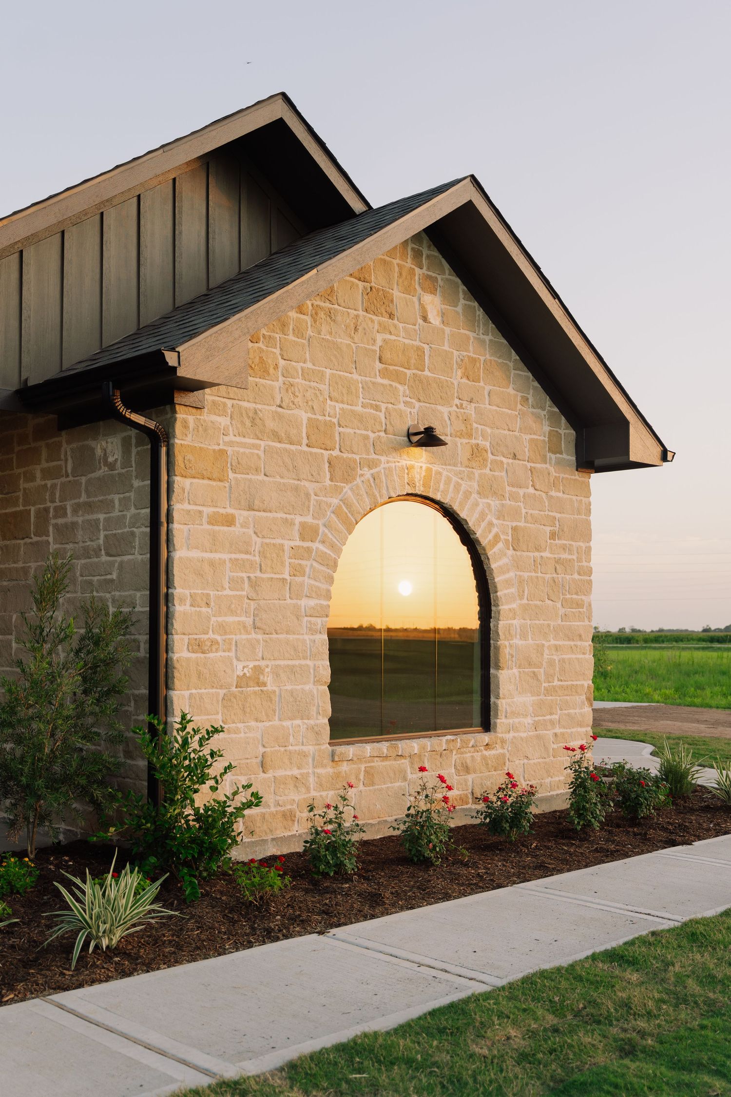 Stone exterior home with arched window reflecting sunset, dark trim, and manicured landscaping at dusk.