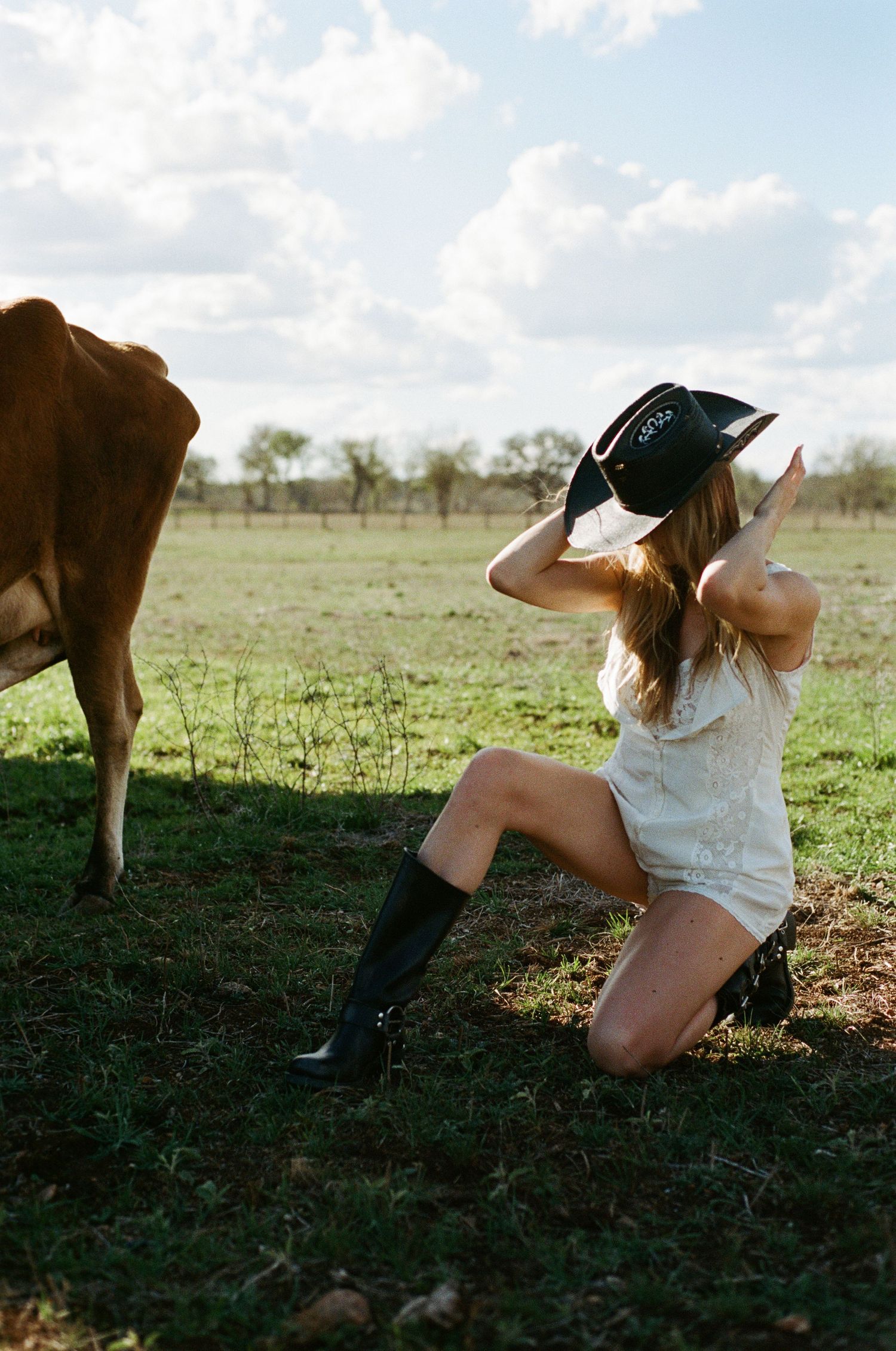 Blonde woman in white dress and cowboy boots posing near a cow in a green pasture.