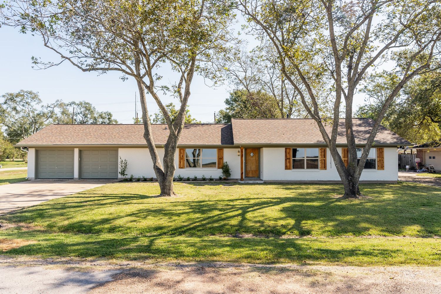Ranch-style home with white exterior, attached garage, wood accents, and mature shade trees on green lawn.