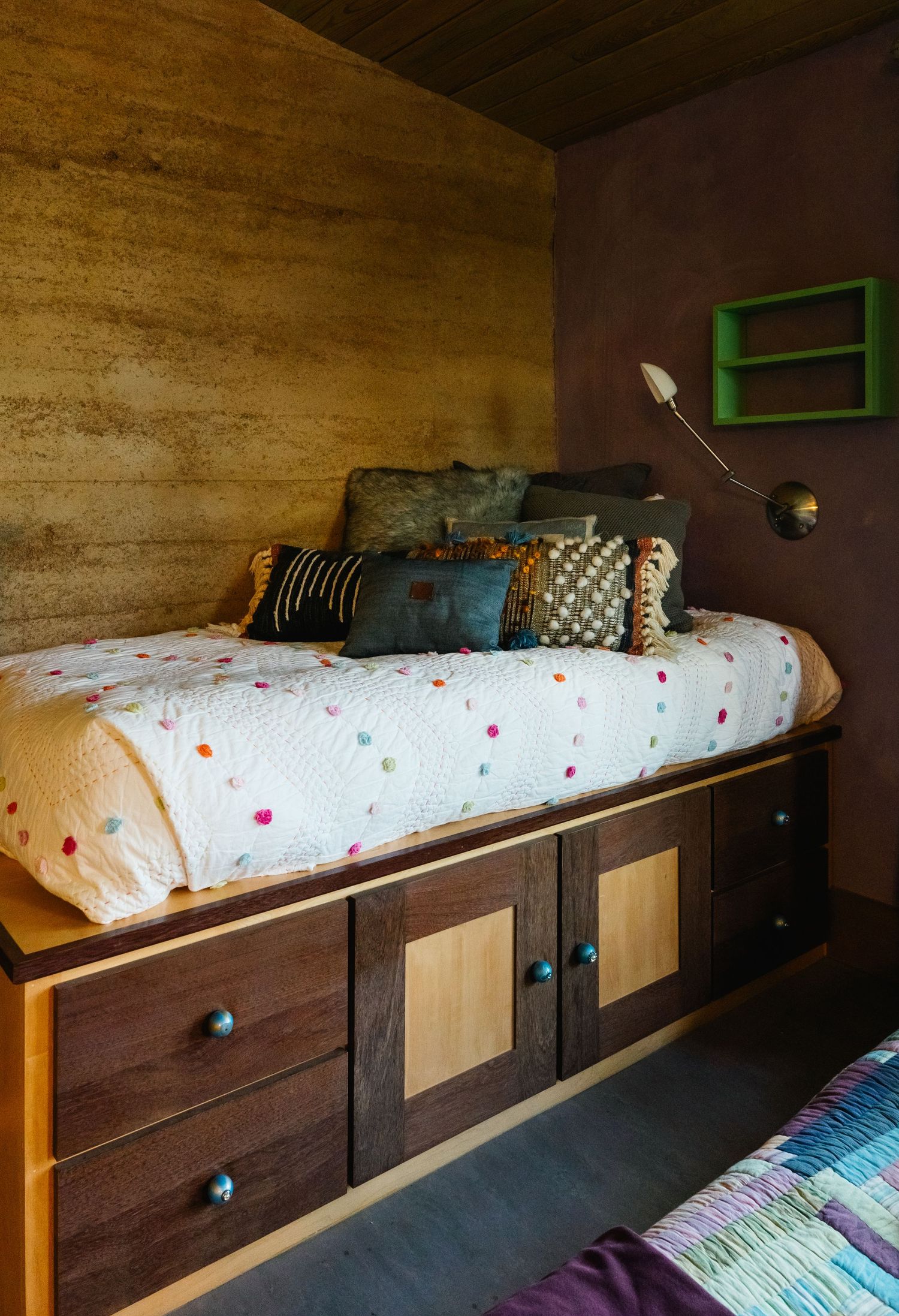 Built-in platform bed with storage drawers, colorful polka dot bedspread, and decorative pillows against wood-paneled wall.