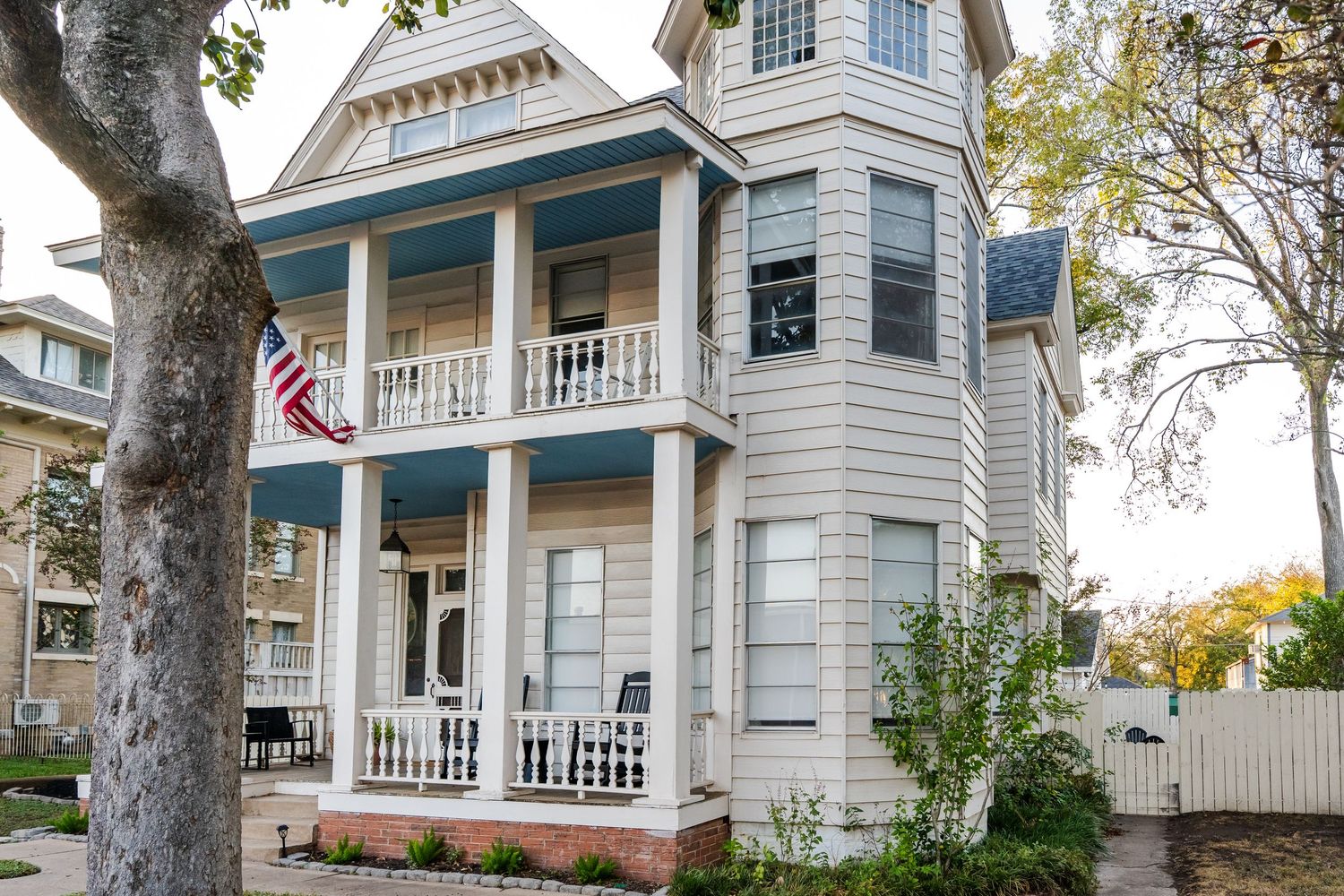 Victorian two-story home with wrap-around porch, blue balcony railings, and American flag on tree-lined street.