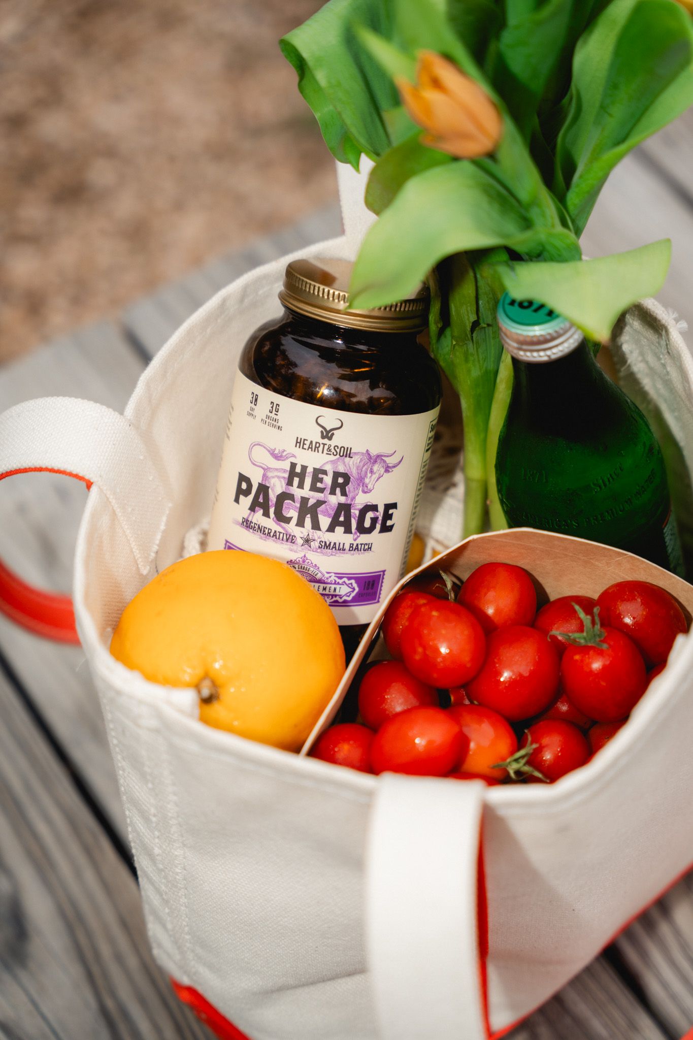 Canvas tote bag with Her Package vitamins, cherry tomatoes, orange, green bottle, and fresh tulips on wooden surface.
