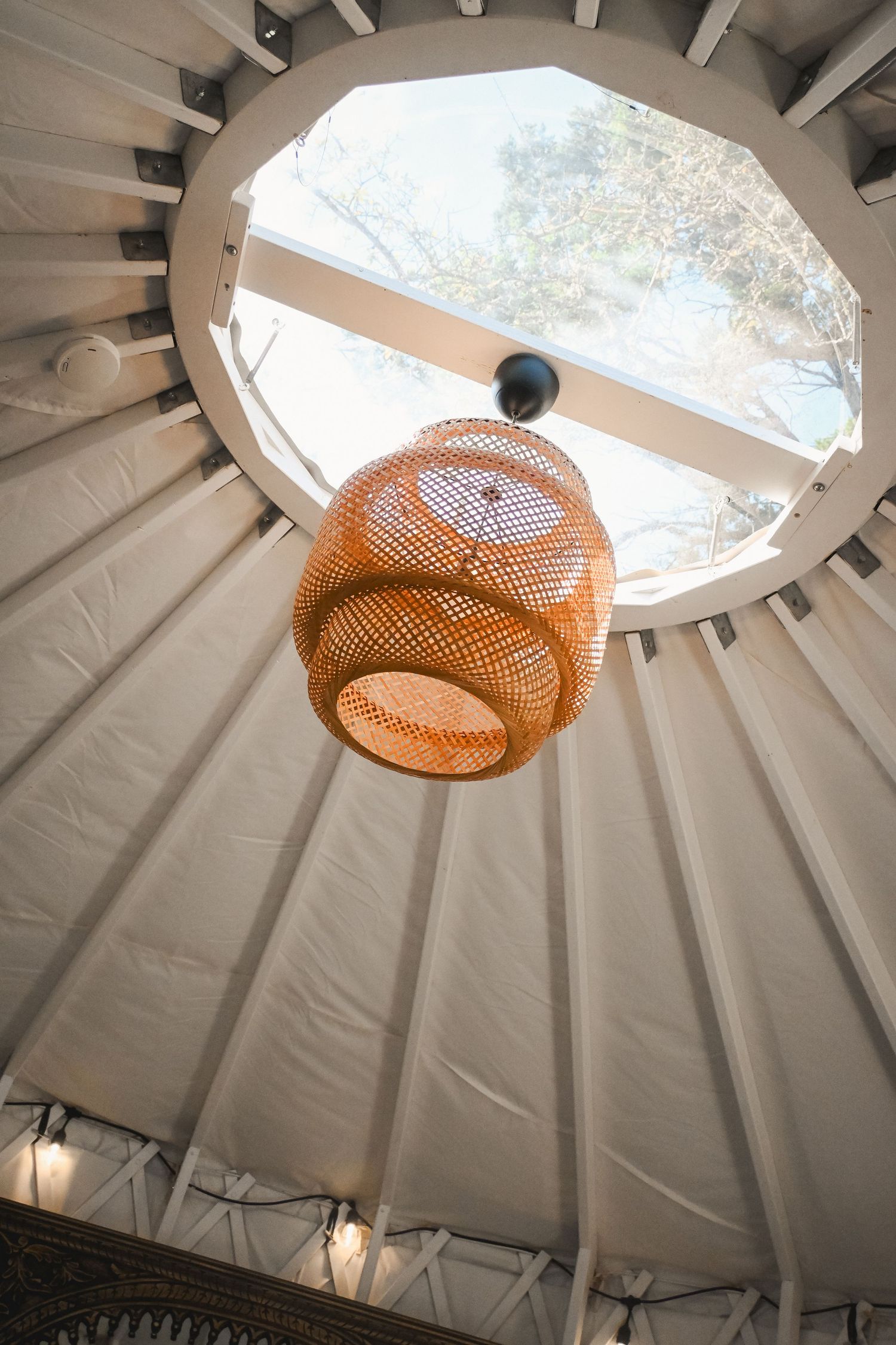 Interior view of a yurt ceiling with a skylight, wicker pendant lamp, and string lights.