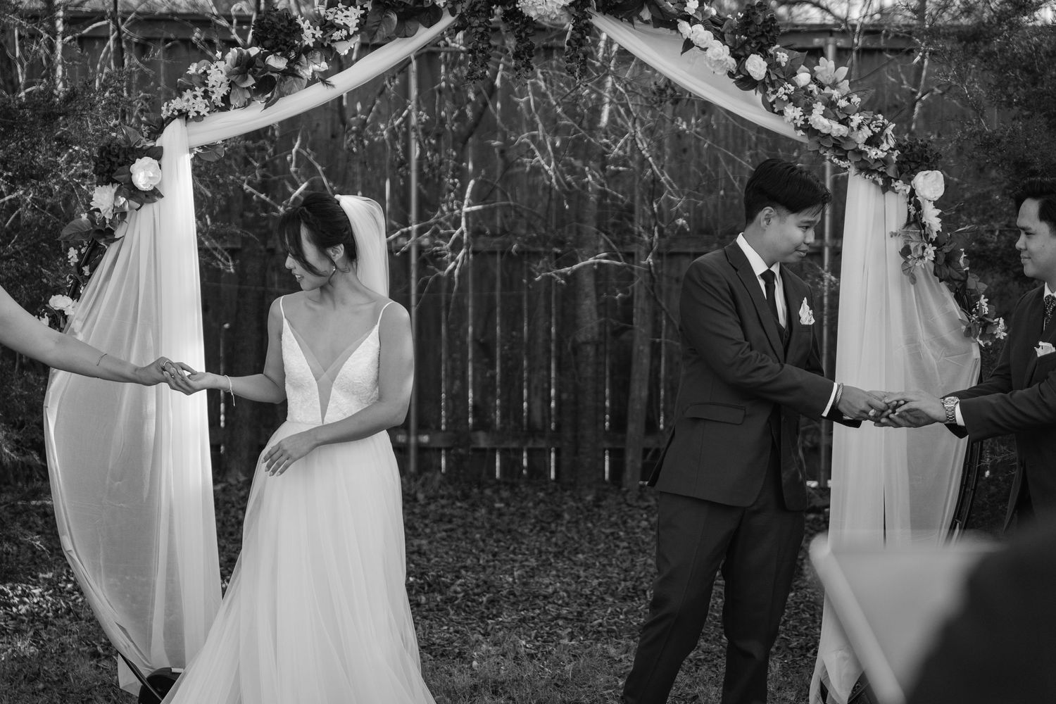 Black and white photo of bride and groom holding hands under floral wedding arch outdoors.