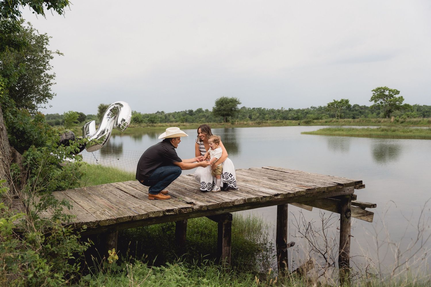Family with toddler enjoying time on a rustic wooden dock by a serene lake surrounded by lush greenery.