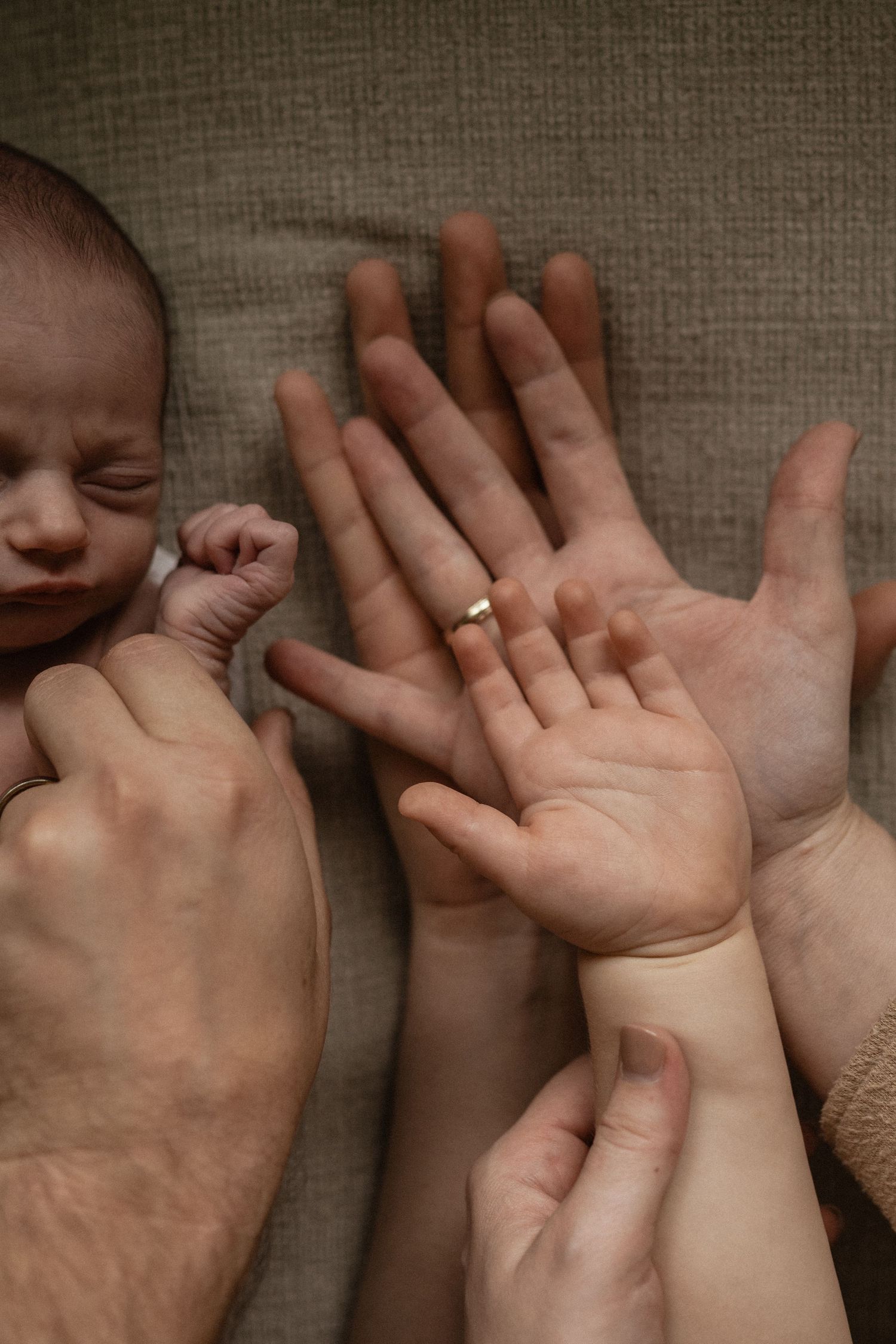 Newborn baby sleeping while parents and sibling hold hands together, showing three generations of family love.